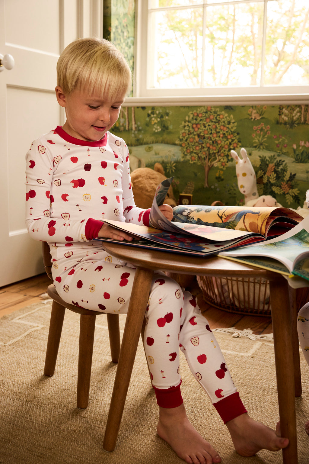 A young child wearing the Kids Long-Long Set in Apple Orchard print pajamas sits on a wooden stool, reading a picture book at a small table in a sunlit, nature-themed room.