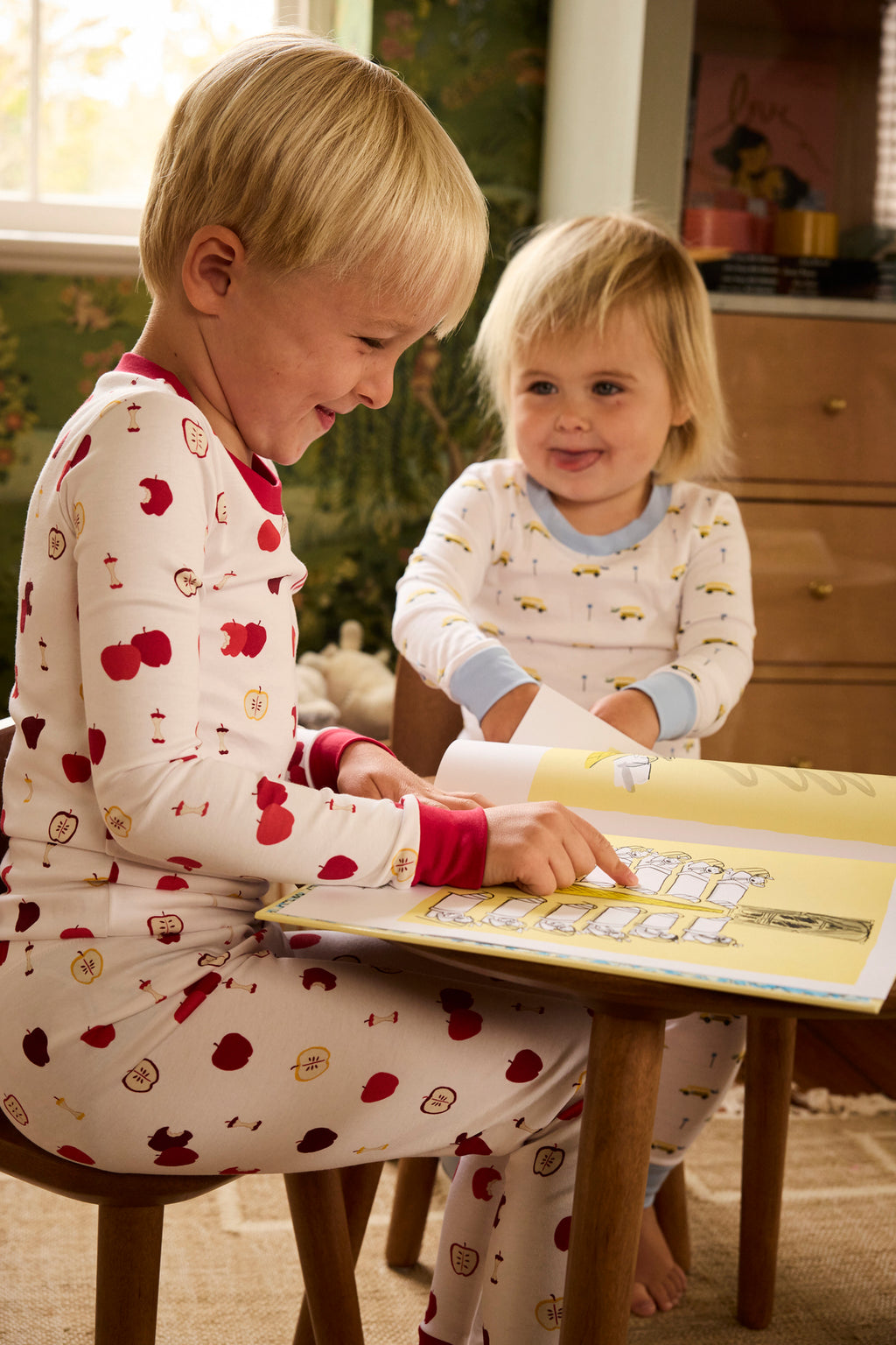 Two young children wearing the Kids Long-Long Set in Apple Orchard Pima cotton pajamas sit happily together indoors, one smiling and pointing at a picture book with an apple print, on wooden chairs in a cozy, warmly lit room.