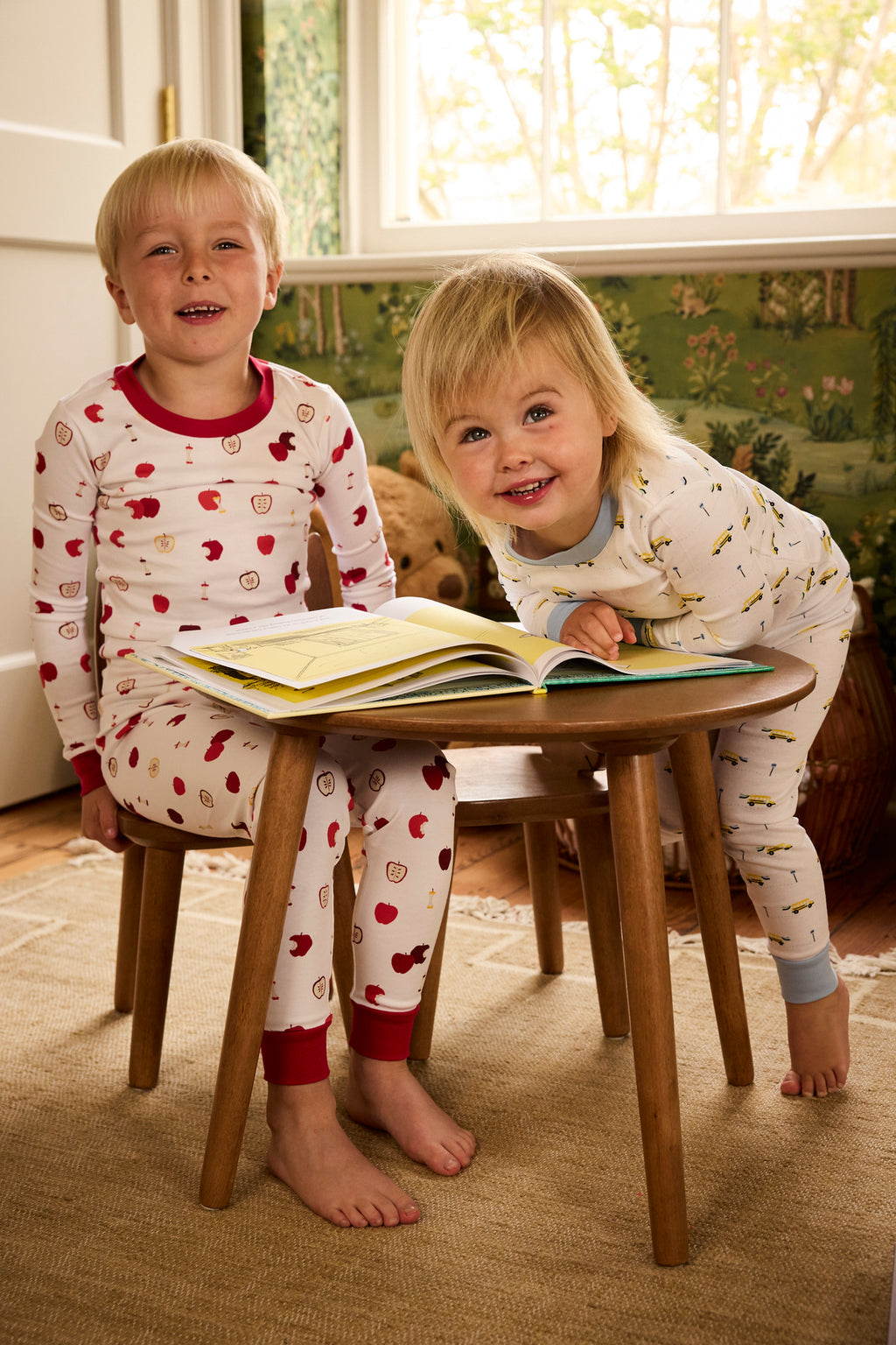 Two young children in Kids Long-Long Set in Apple Orchard pajamas smile as they gather around a small wooden table with an open book in a cozy, sunlit room, one seated and the other leaning on the table, both looking happy and playful.