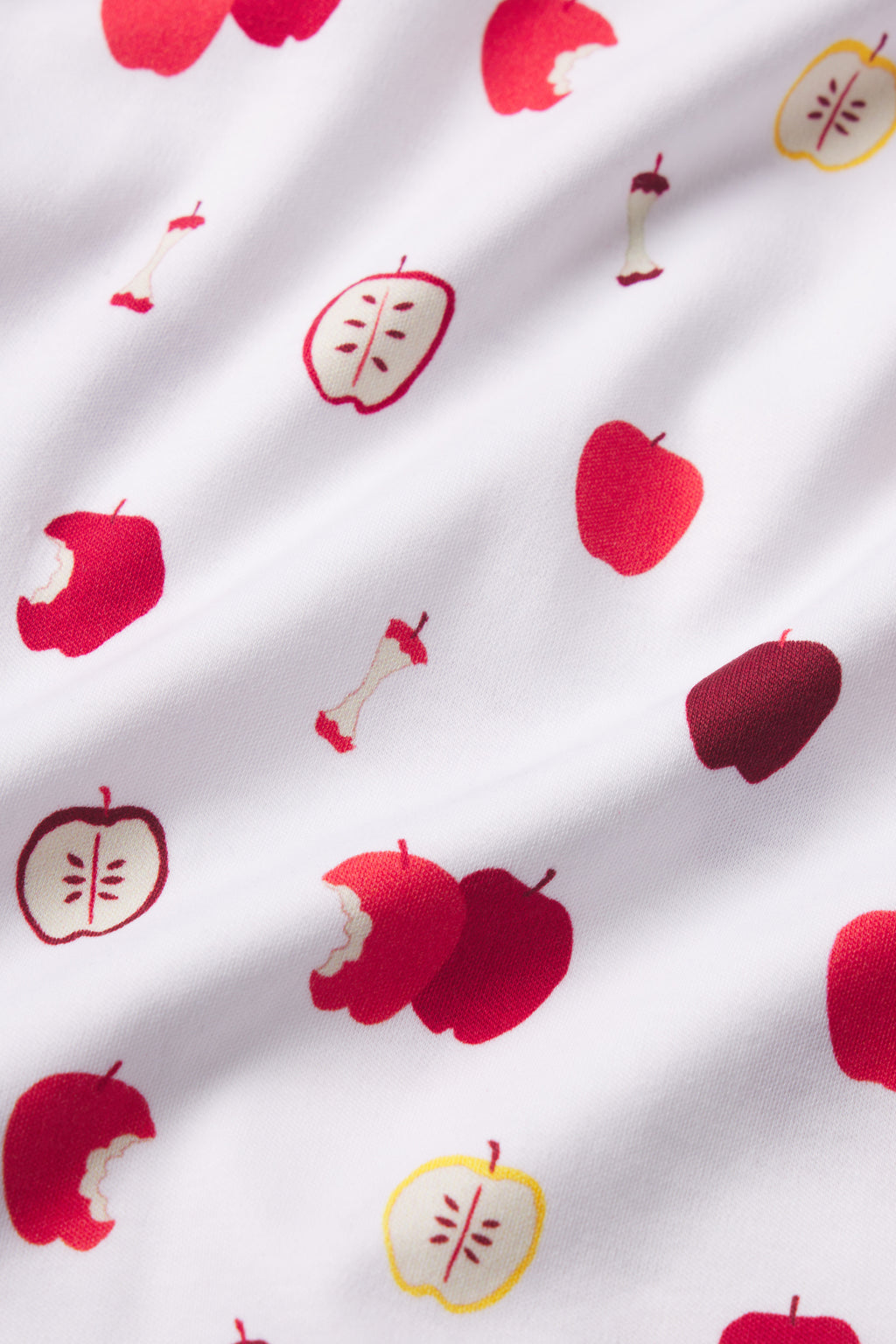 The Kids Shorts Set in Apple Orchard features a softly wrinkled white Pima cotton fabric adorned with red and yellow apples—whole, bitten, and cored—for a playful and charming look.