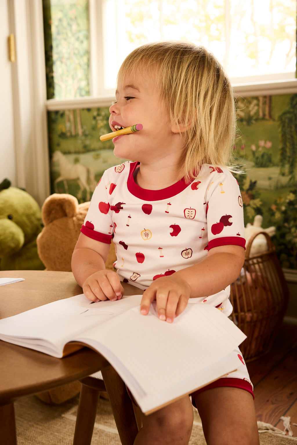 A young child in the Kids Shorts Set in Apple Orchard sits at a small desk, smiling with a pencil in their mouth, holding an open notebook amid stuffed animals and nature-themed wallpaper.