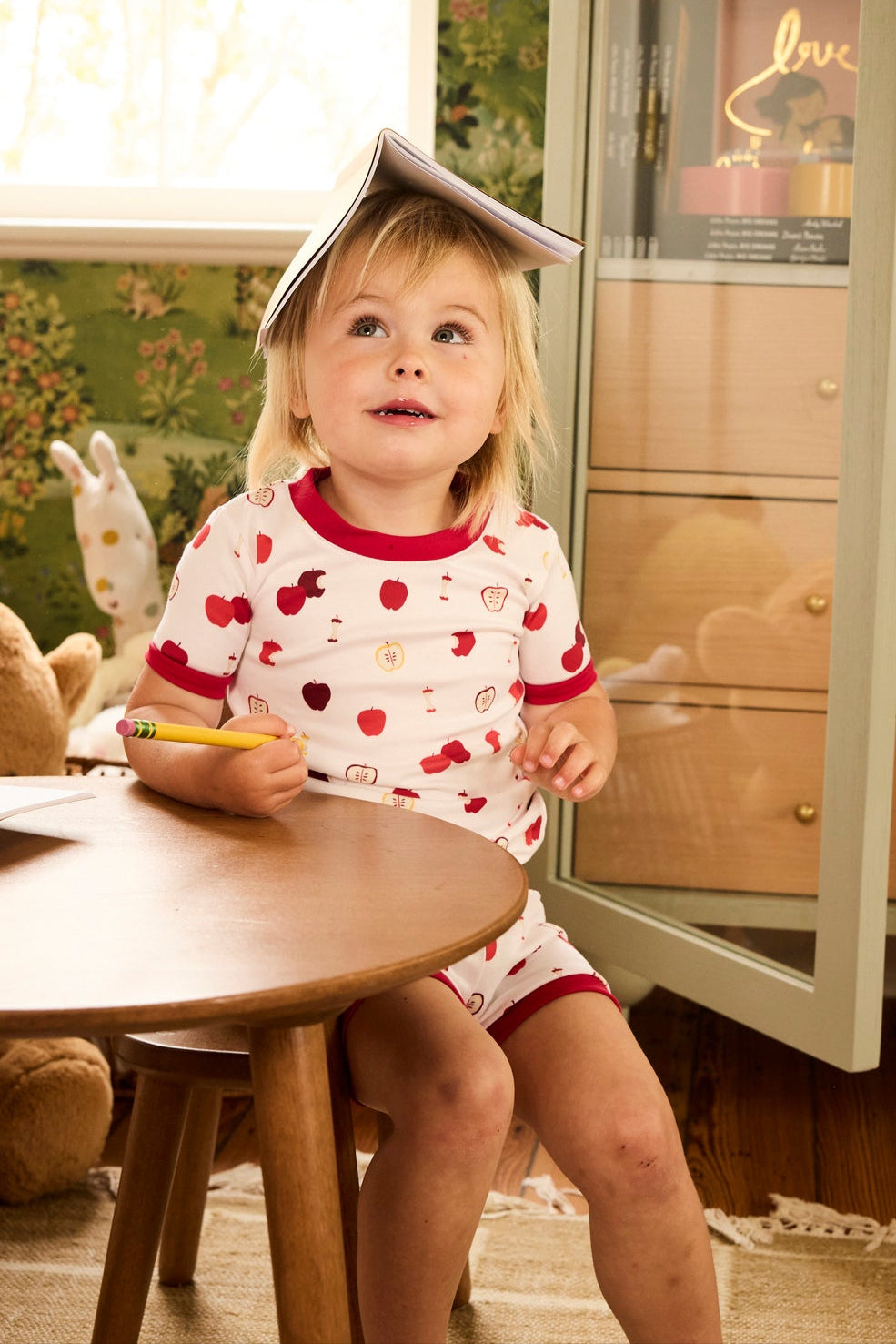 A young child wearing the Kids Shorts Set in Apple Orchard sits at a small wooden table, holding a pencil with a notebook on their head. Toys, a bunny plush, and green floral wallpaper are in the background.