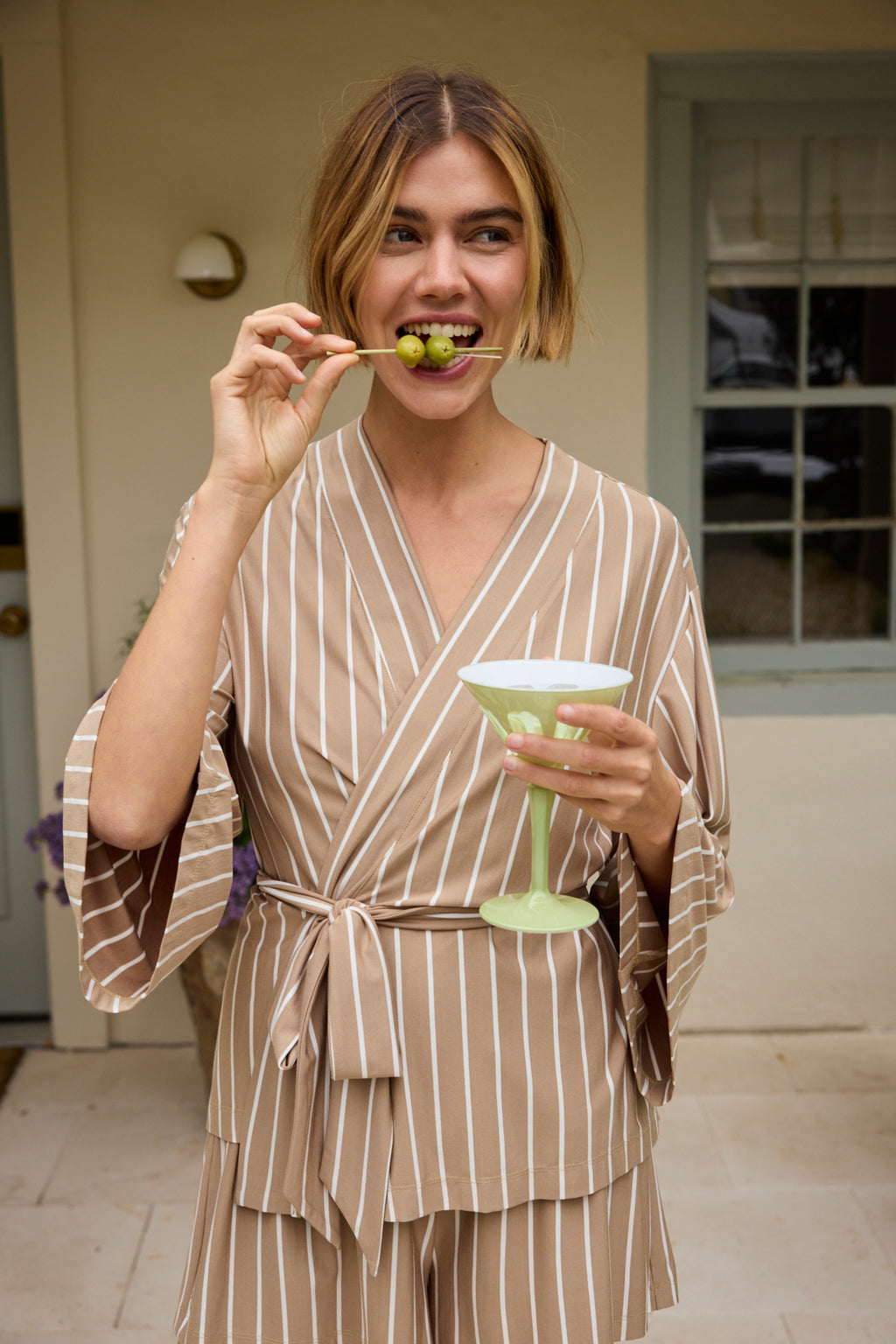 Smiling, a woman in the DreamModal Kimono Shorts Bundle in Sandstone Vertical Stripe stands outside by a window and beige wall, holding a green cocktail and enjoying grapes from a stick.