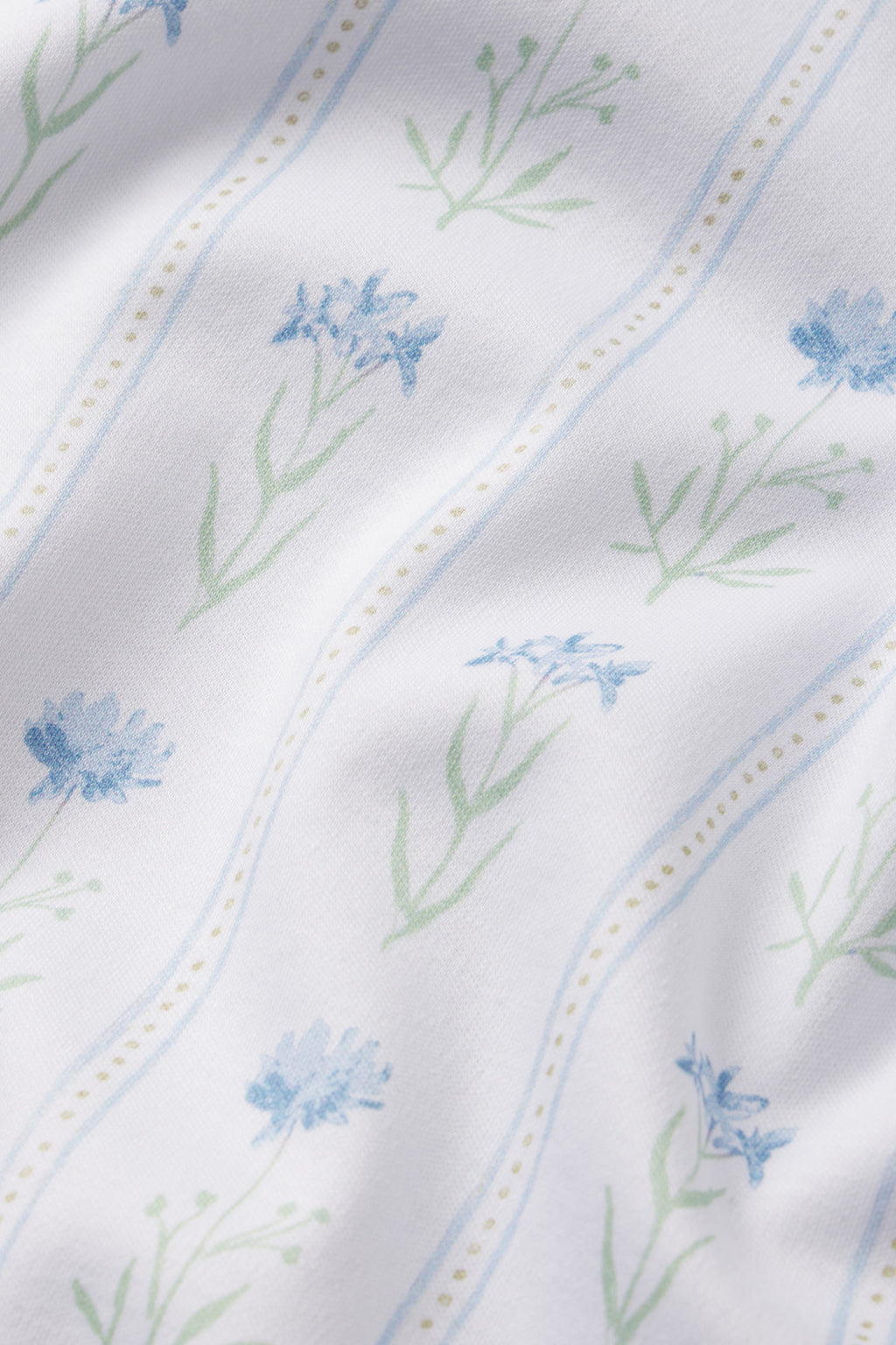 The Pima Bundle in Serene Blue Watercolor Stems features white Pima cotton with blue watercolor floral designs, green stems, and thin blue and dotted yellow vertical lines, accented by soft folds and shadows on the fabric surface.
