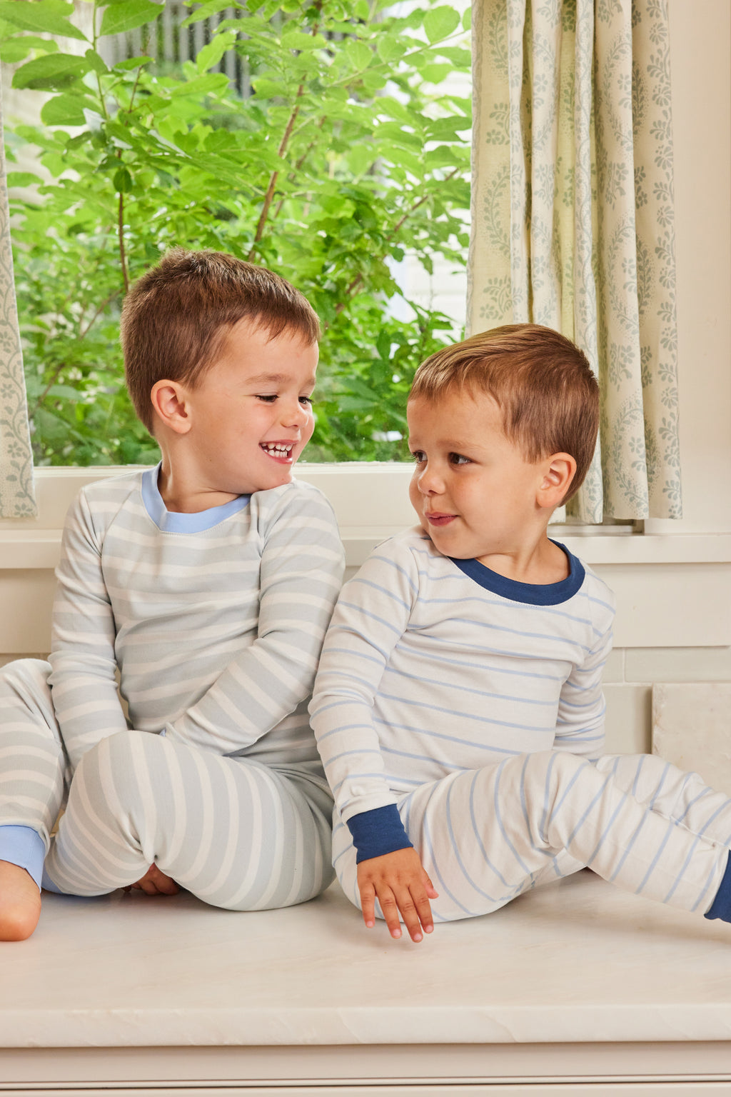 Two young boys in matching Kids Long-Long Set in Seaside pajamas sit on a windowsill, smiling at each other with green foliage visible through the window behind them.