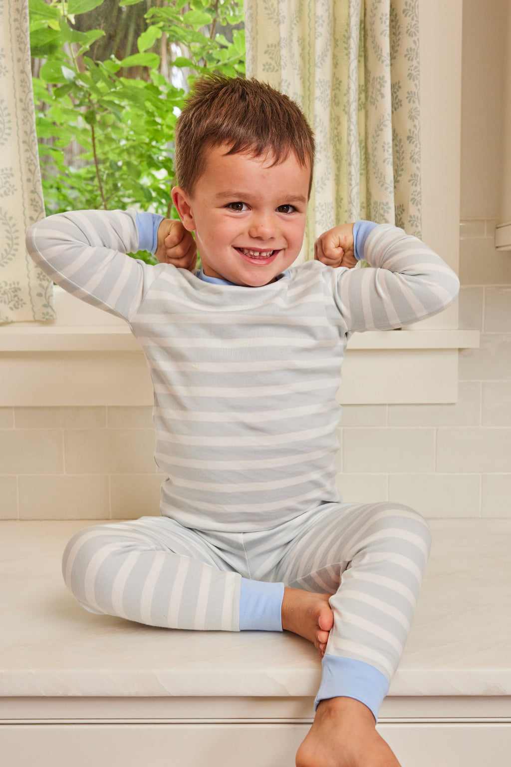 A smiling young boy wears the Kids Long-Long Set in Seaside as he sits on a windowsill, flexing his arms with green leaves visible outside the window behind him.