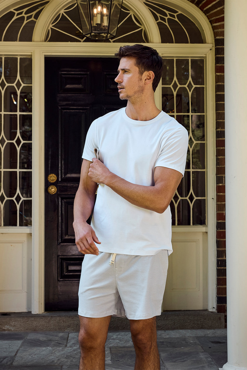 A man in a white t-shirt and Mens Pima Sleep Shorts in Slate Bar Stripe stands outside before a dark wooden door with decorative glass, looking to his left with one arm slightly raised.