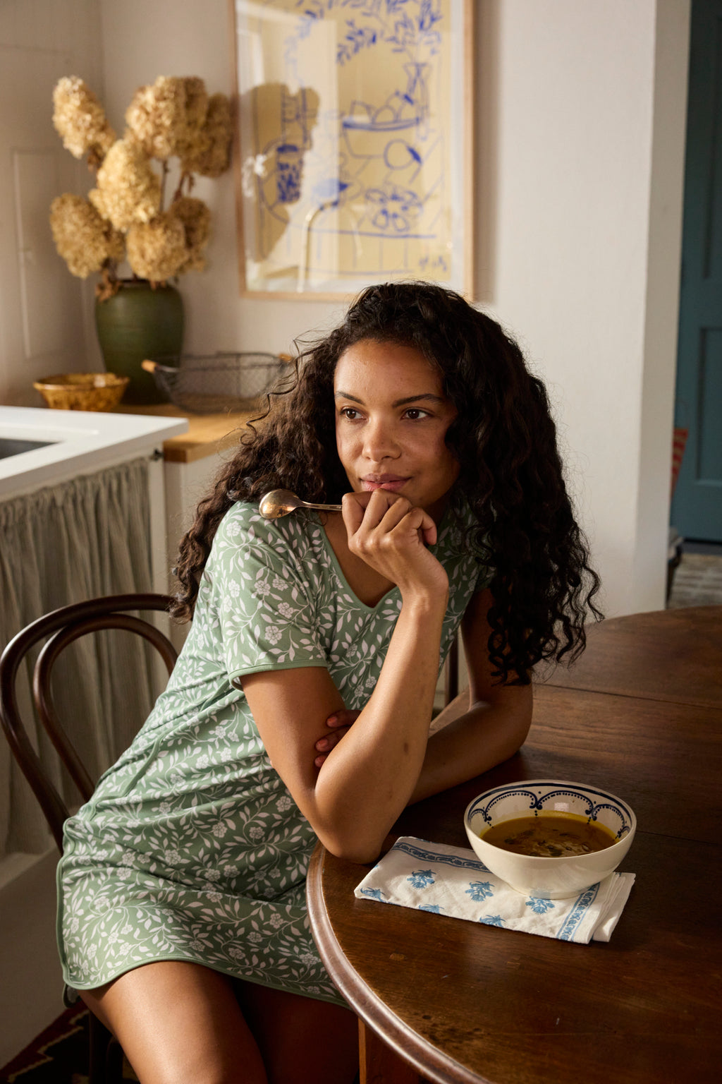 A woman in a Pima Nightgown in Granite Green Cecil Floral sits at a wooden table, thoughtfully tasting soup. A napkin rests nearby, with dried flowers and artwork accenting the kitchen.
