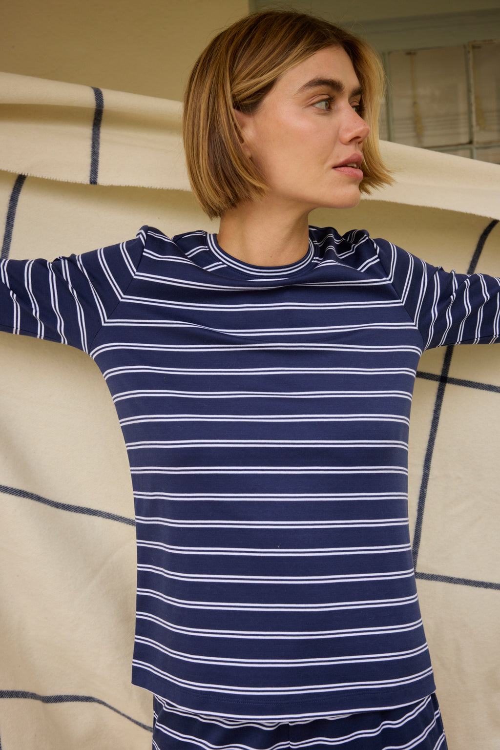 A person wearing the Pima Crew Long Sleeve Bundle in English Navy Tidal Stripe stands with arms outstretched in front of a beige blanket with blue stripes.