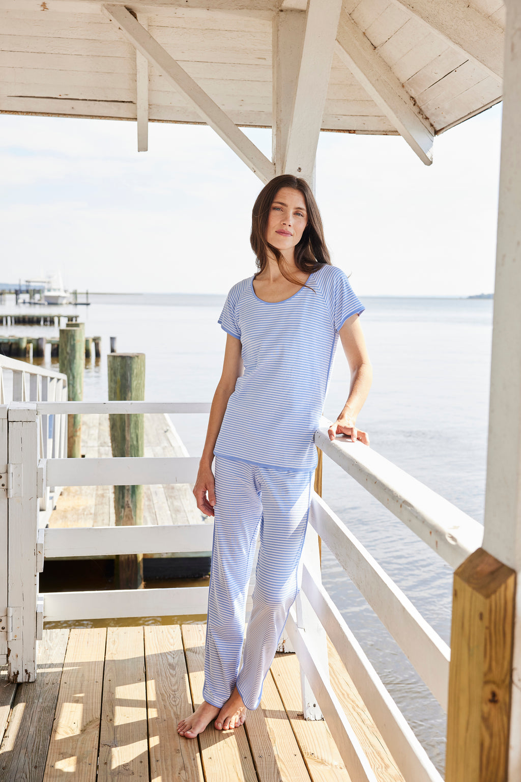 Wearing the Pima Short-Long Set in Hydrangea, a woman stands on a wooden deck overlooking the water, framed by a white roof and pier posts. The soft Hydrangea hues enhance the tranquil atmosphere.