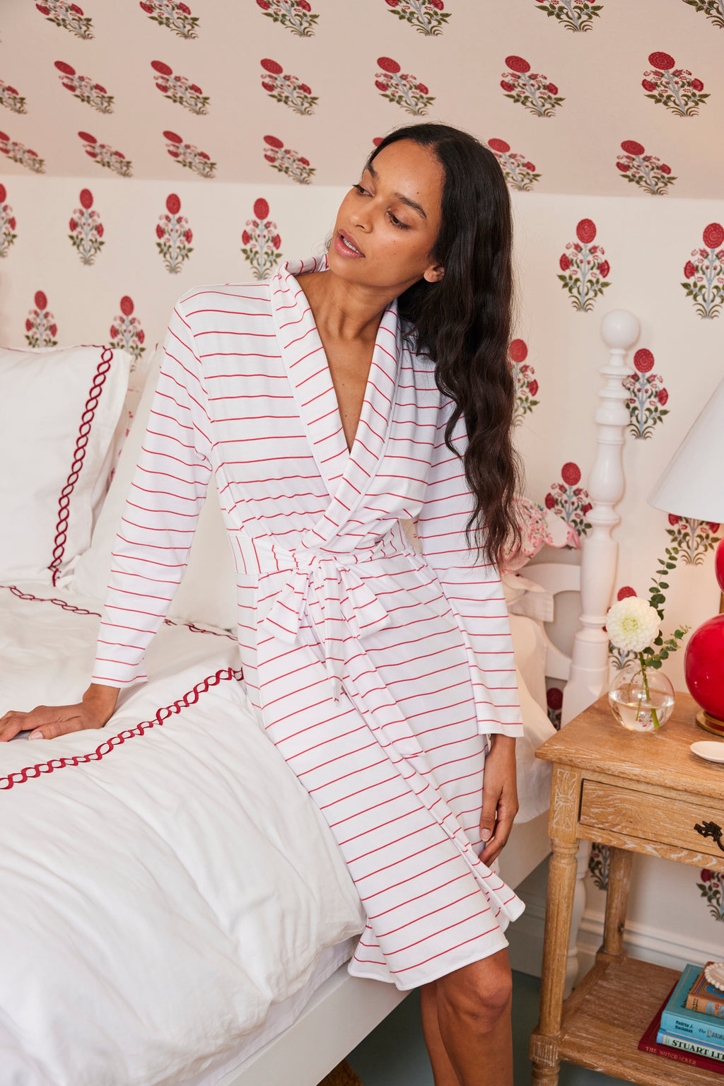 A woman sits on a bed wearing the Pima Robe in Geranium Boating Stripe. The room has floral wallpaper, a white headboard, and a wooden nightstand topped with a lamp and flowers.
