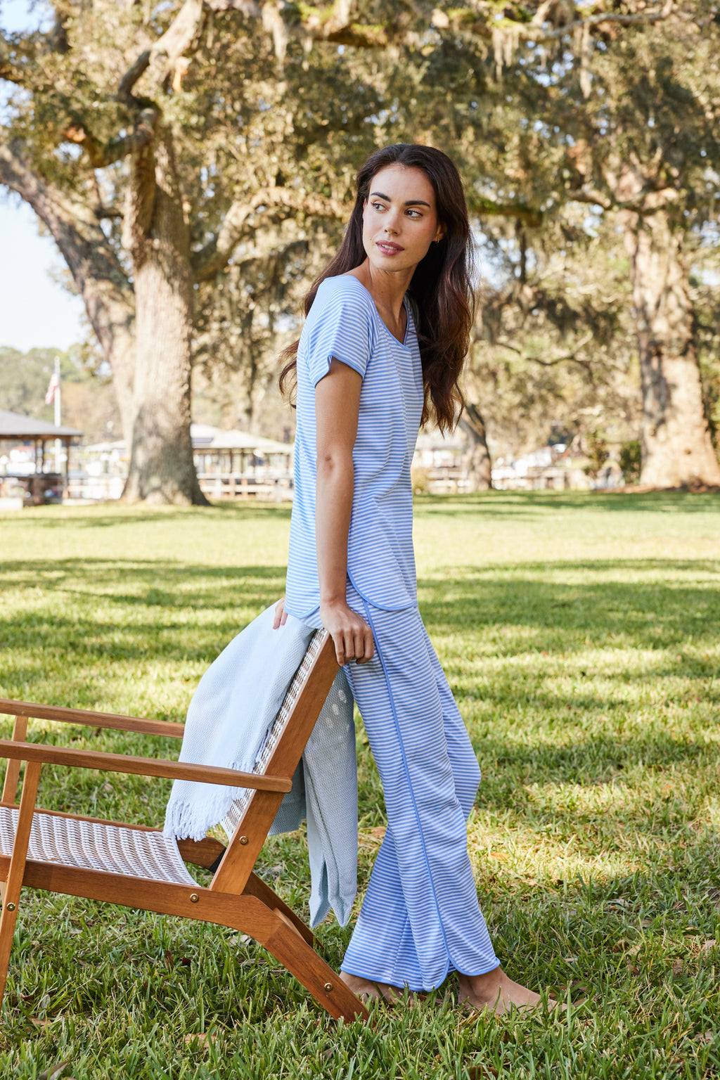 A woman wearing the Pima Short-Long Set in Hydrangea sits on a wooden chairs arm on a grassy lawn, holding a white blanket, with sunlight and large trees in the background.