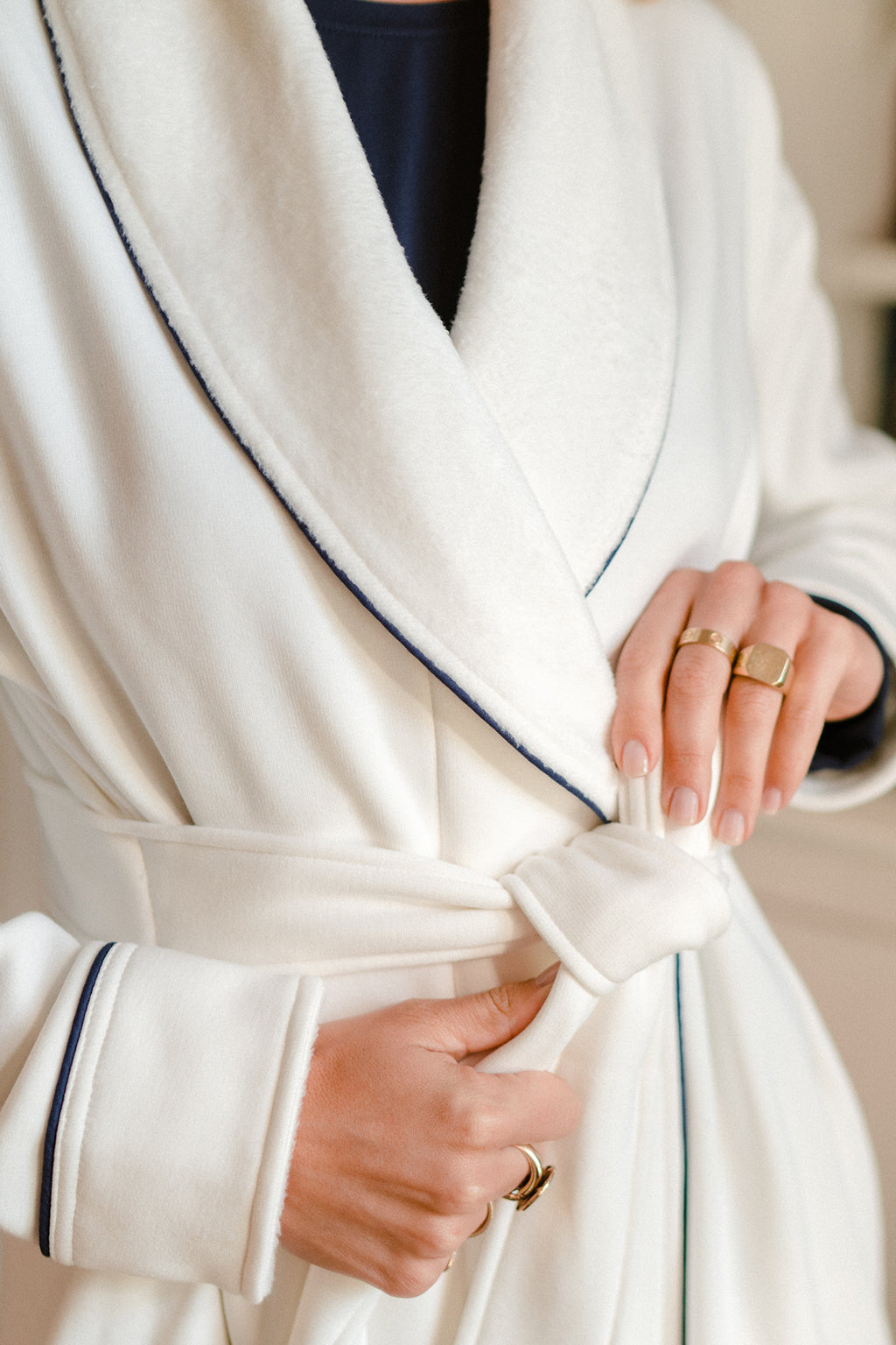 A person ties the belt of a Cozy Robe in Navy, crafted from soft cotton jersey with dark piping, showcasing neatly manicured nails and gold rings on their fingers.