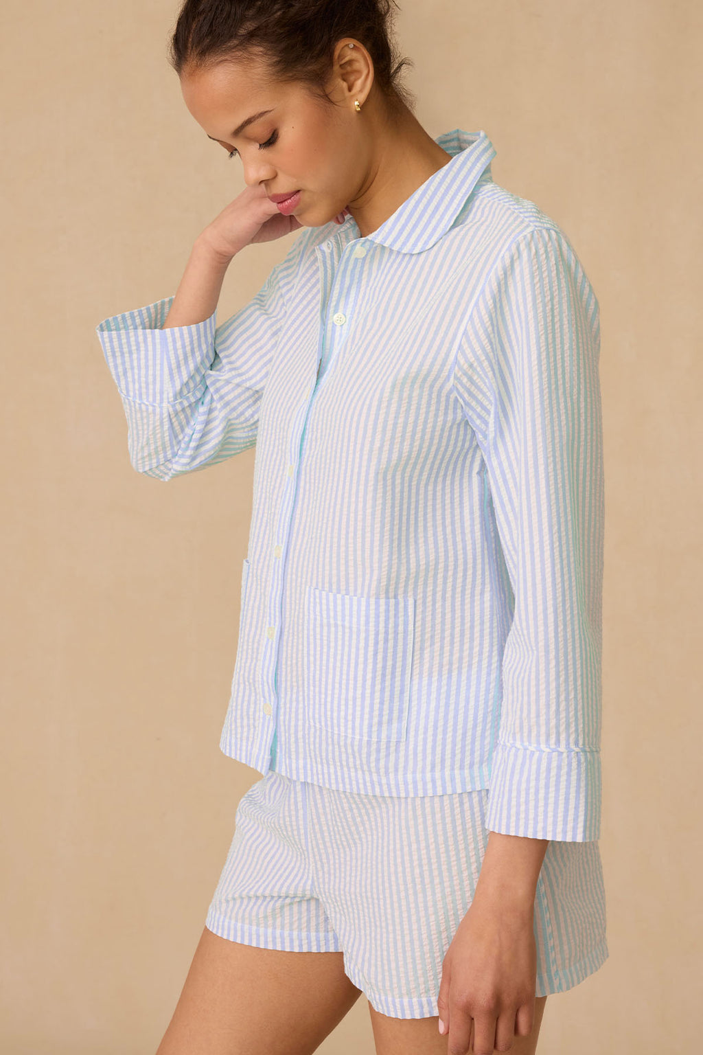 A woman stands against a beige background, wearing the Georgie Pocket Shorts Set in Salt Spray Seersucker—a light blue and white striped Pima cotton pajama set with a collared button-up top and matching shorts. She tilts her head slightly down.
