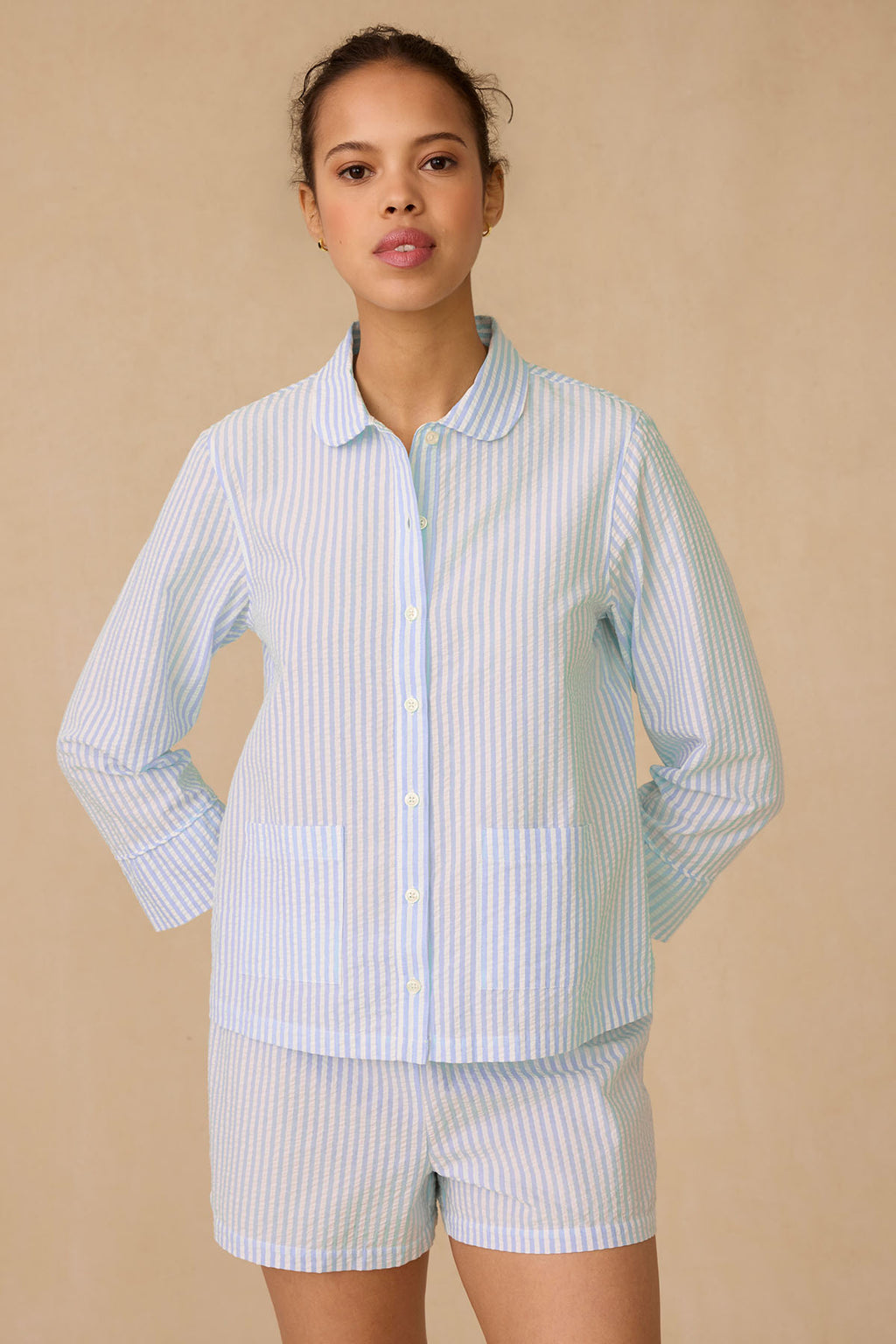 A woman poses against a beige background in the Georgie Pocket Shorts Set in Salt Spray Seersucker, which includes a light blue and white striped button-up long-sleeve top and matching shorts. She stands with her hands behind her back, facing the camera.