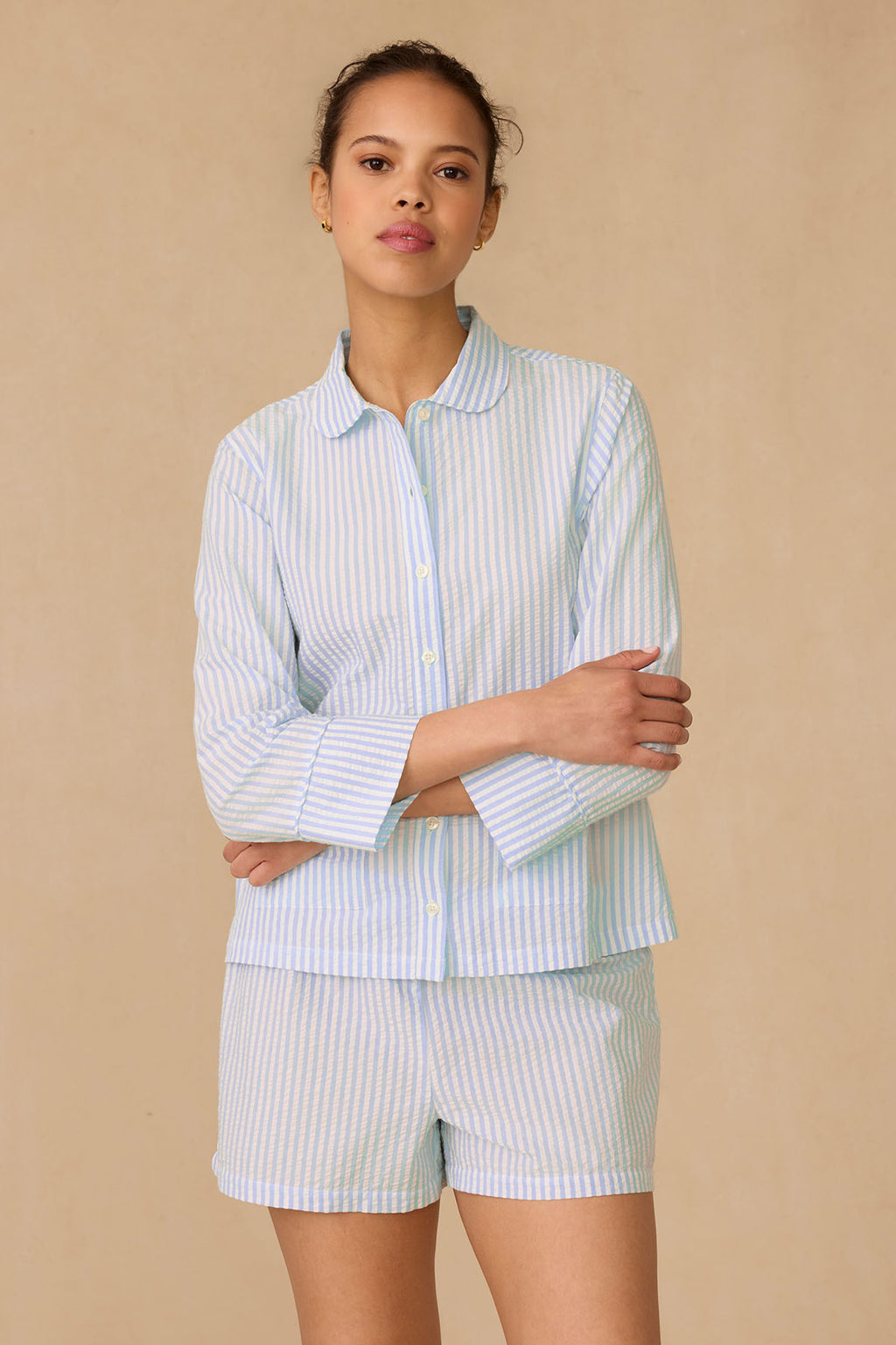 Against a plain beige background, the model wears the Georgie Pocket Shorts Set in Salt Spray Seersucker—light blue and white striped button-up top with matching shorts—standing with arms crossed and a neutral expression.