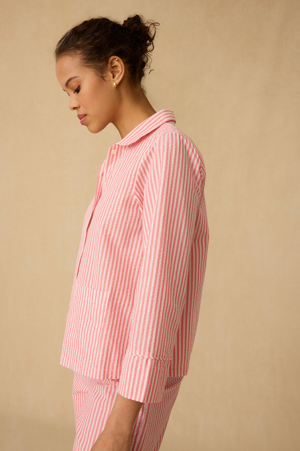 Wearing the Georgie Pocket Pants Set in Gelato Seersucker, she stands sideways before a beige background, gazing down in calm elegance. The pink and white striped set features a long-sleeve top and matching pants for effortless style.