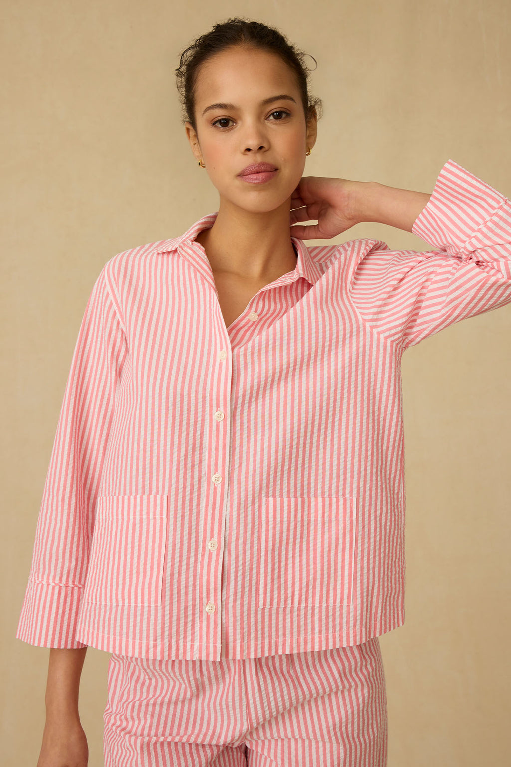 A woman models the Georgie Pocket Pants Set in Gelato Seersucker, featuring pink and white stripes, standing against a beige background with one hand behind her neck.