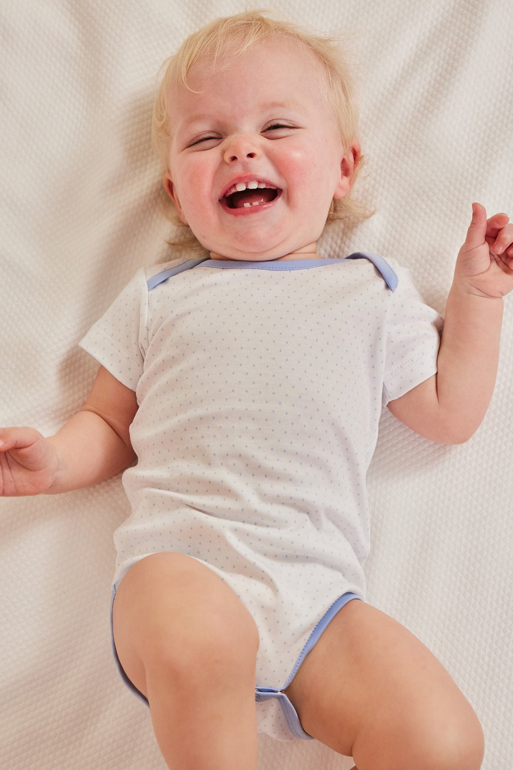 A smiling baby lies on a white blanket, wearing the Baby Bodysuit Bundle in French Blue, made from 100% Pima cotton with small dot patterns.