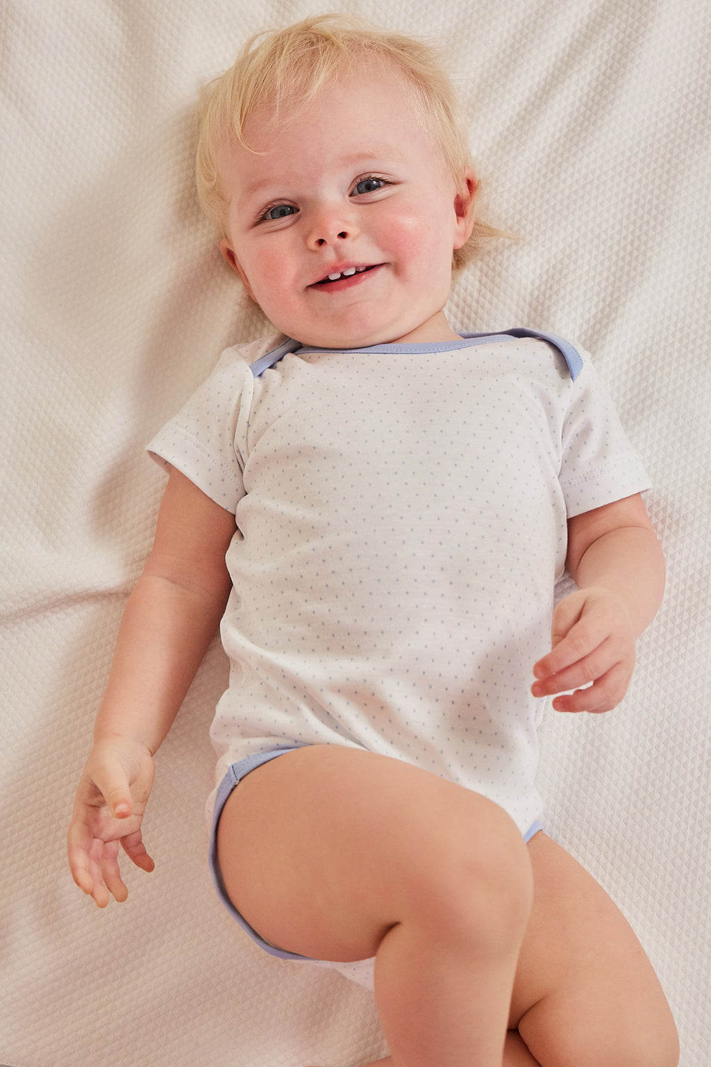 A cheerful toddler lies on a light blanket, wearing the Baby Bodysuit Bundle in French Blue—100% Pima cotton with blue dot details—and looks up with a smile.