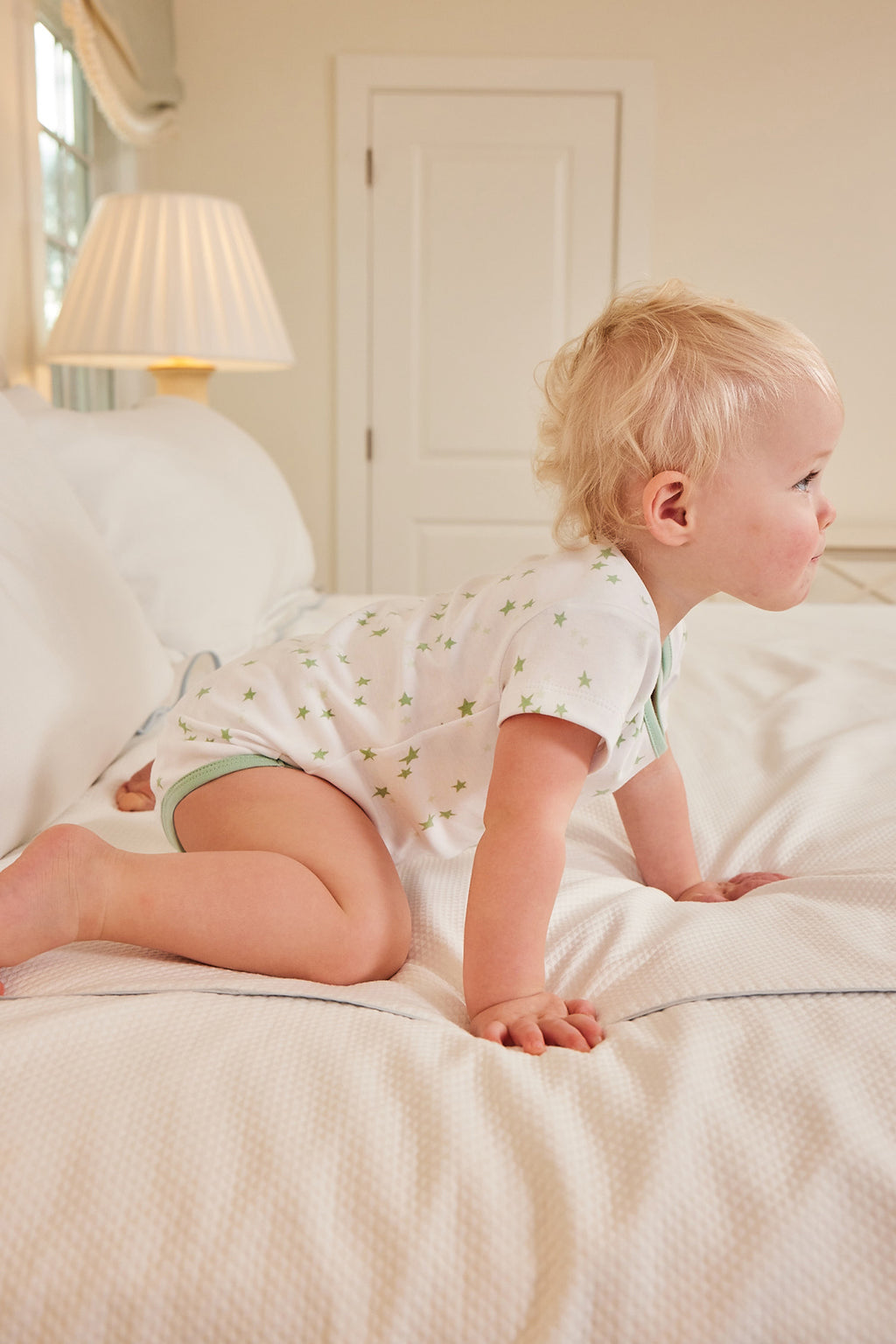 A blonde toddler in a Baby Bodysuit Bundle in Parisian Green crawls on a white bed in a softly lit bedroom, with a lamp and window in the background.