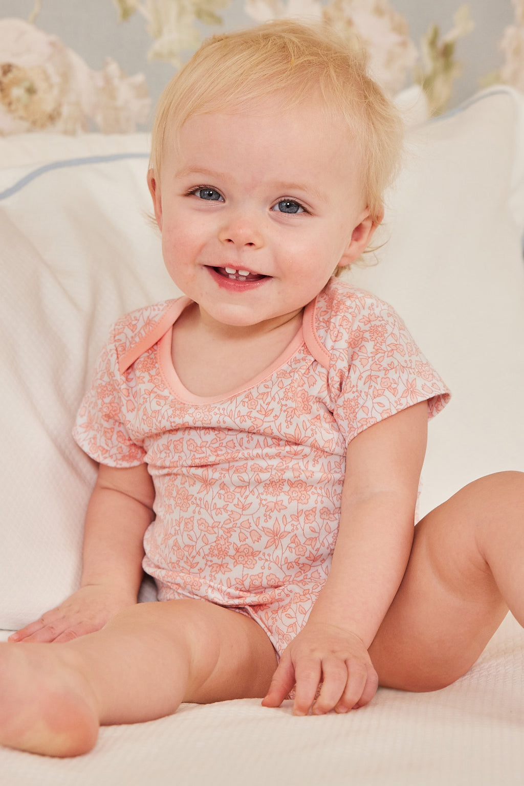 A smiling baby sits on a bed, wearing the Baby Bodysuit Bundle in English Rose. The Pima bodysuit has a light pink floral pattern and is made from 100% Pima cotton, with soft neutral pillows and floral wallpaper in the background.