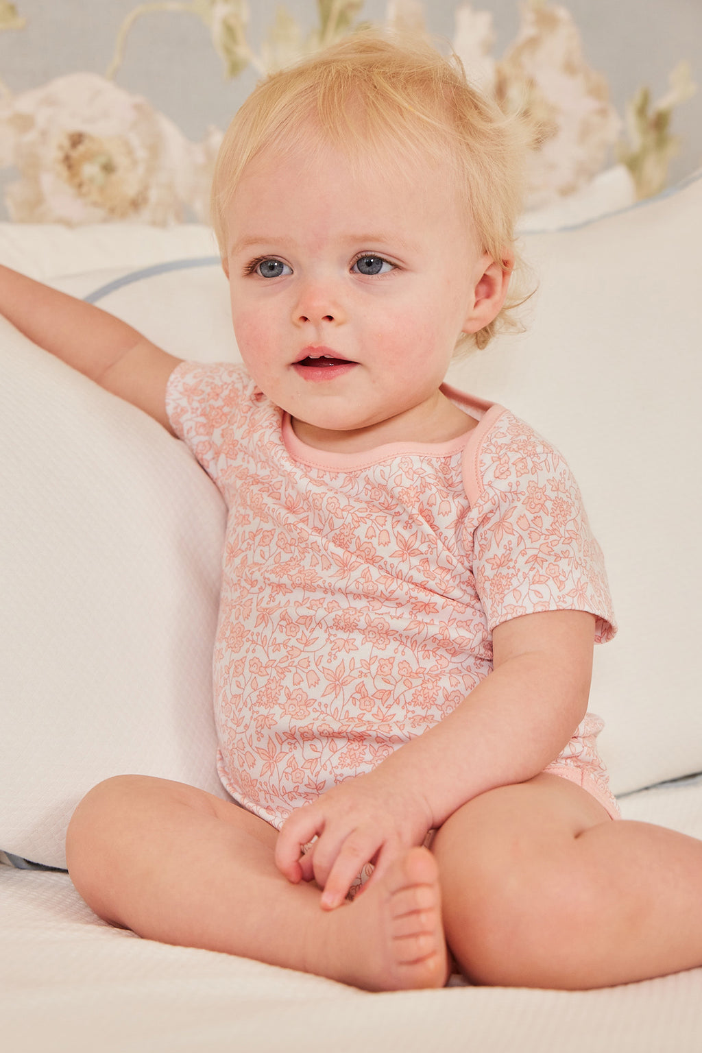 A blonde baby in the Baby Bodysuit Bundle in English Rose sits on a white bed, resting one arm on a pillow and gazing slightly to the side.