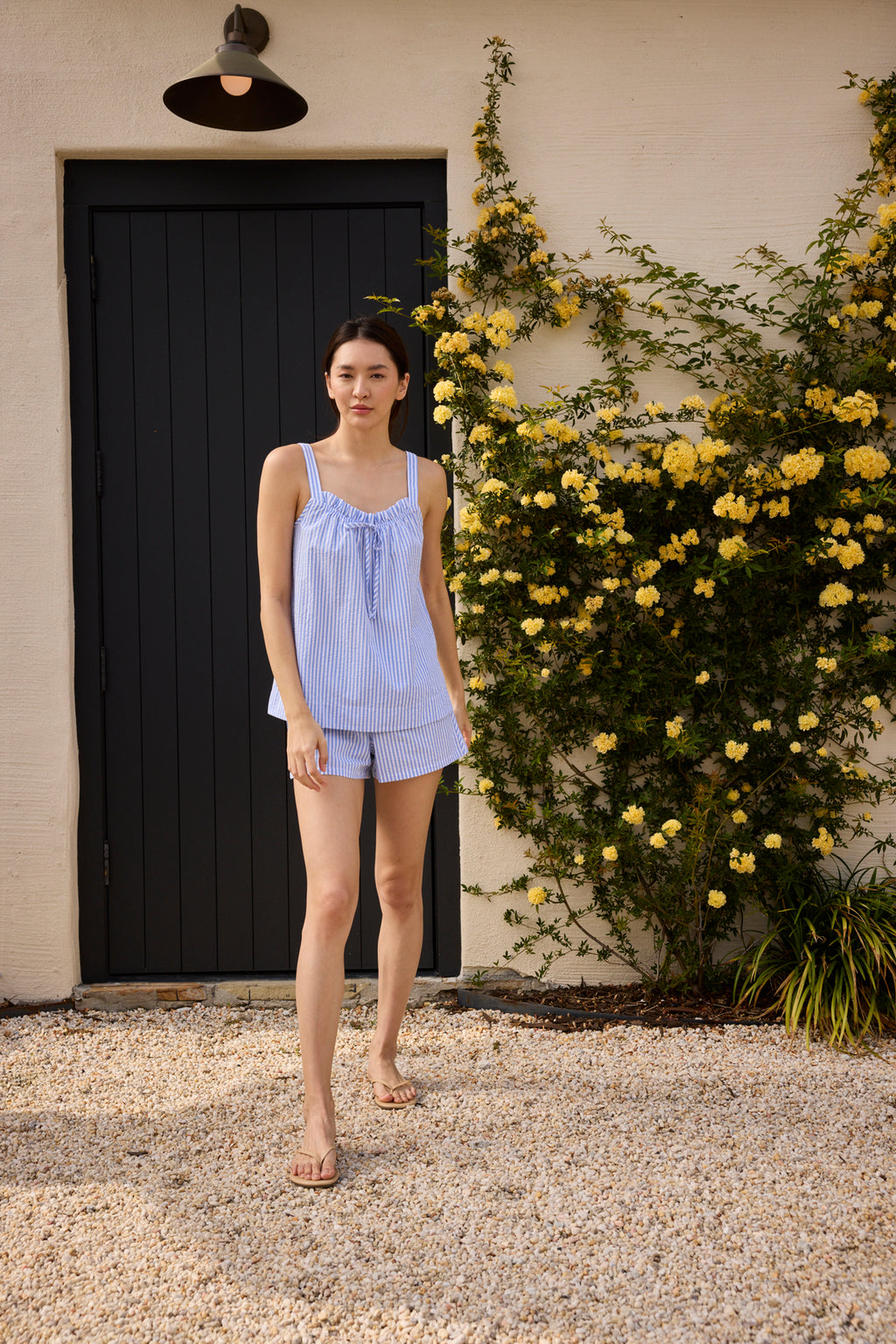 A woman wearing the Alice Shorts Set in Blue Seersucker stands on a gravel path, with yellow flowers climbing a wall and a black door in the background.