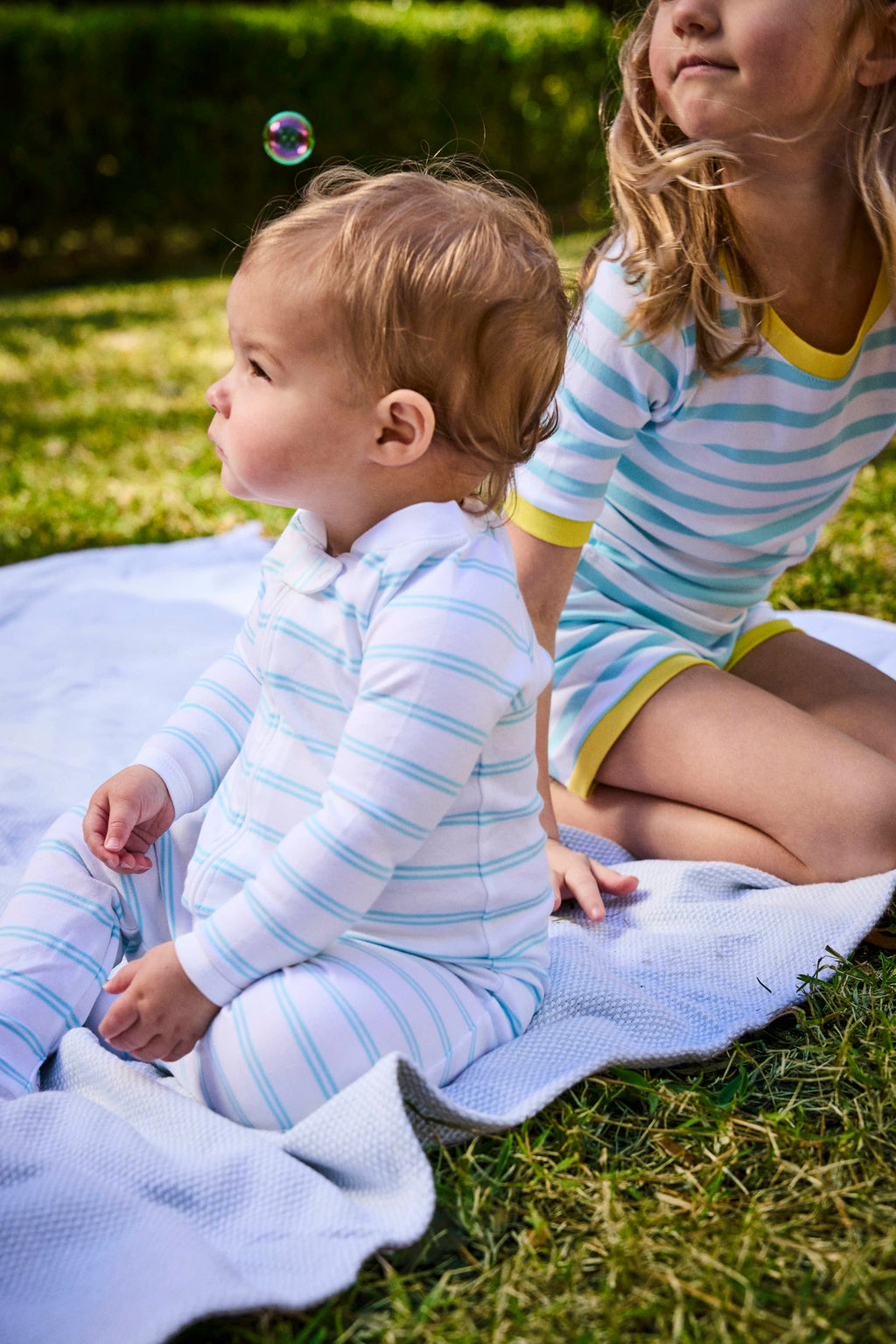 A baby wearing the Baby Sleeper in Beach Club Tidal Stripe sits on a blanket outdoors next to an older child in a matching outfit, surrounded by grass and greenery, with a bubble floating above the babys head.