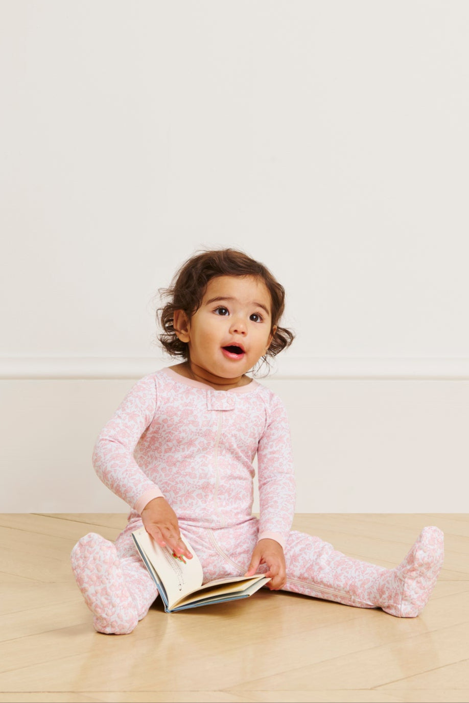 A toddler in a Baby Sleeper in English Rose Garden Floral sits on a wooden floor with legs outstretched, holding an open book and looking up with a surprised expression.