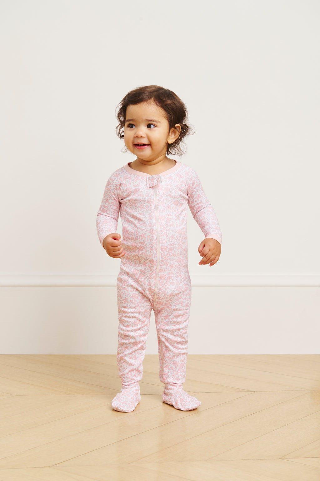 A toddler smiles on a light wood floor, wearing the Baby Sleeper in English Rose Garden Floral—an ultra-soft, long-sleeve 100% Pima cotton outfit with a subtle white floral pattern. A plain, light-colored wall forms the background.