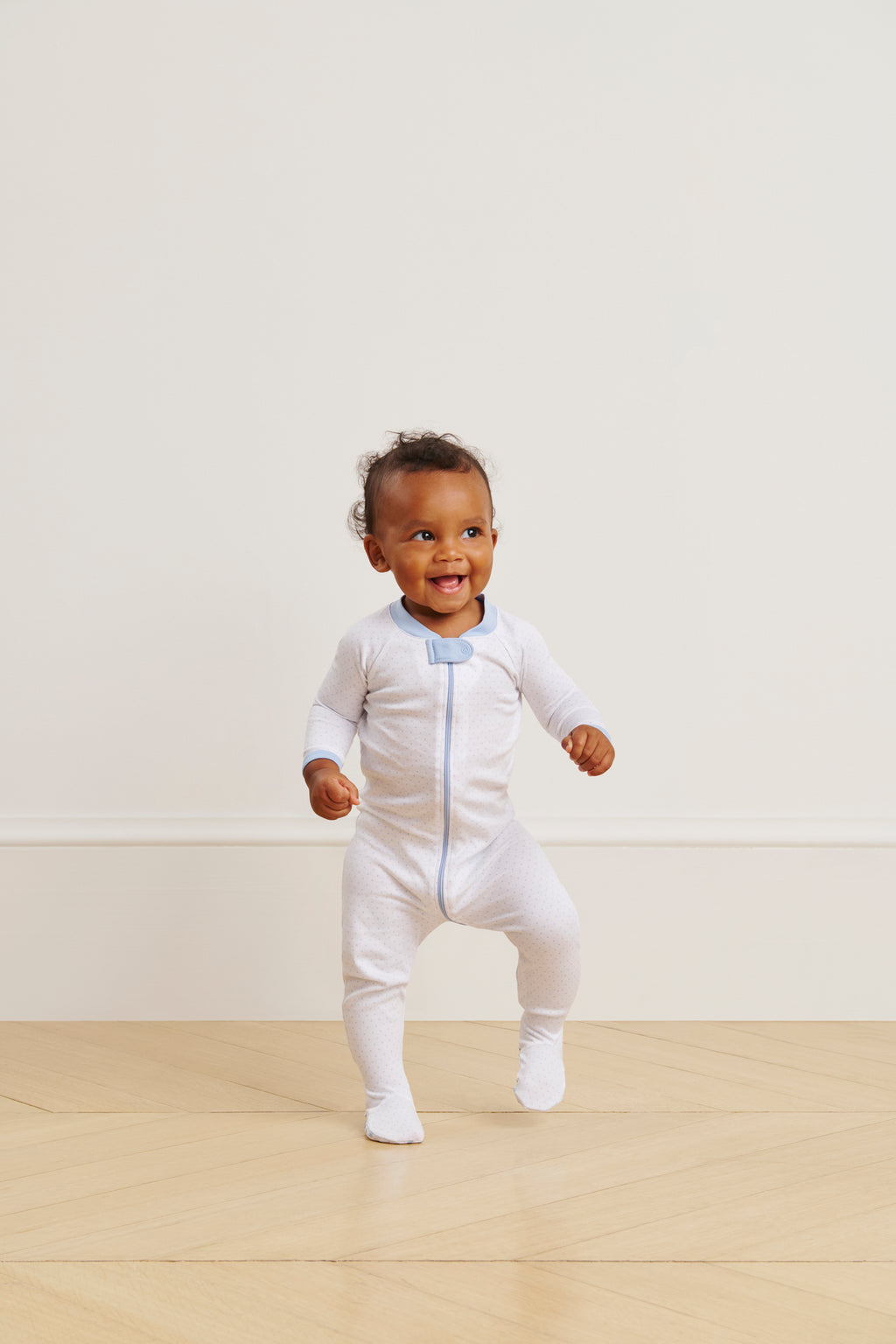 A smiling baby in the Baby Sleeper in French Blue Pindot stands and takes a step on a light wooden floor against a plain, light-colored wall.