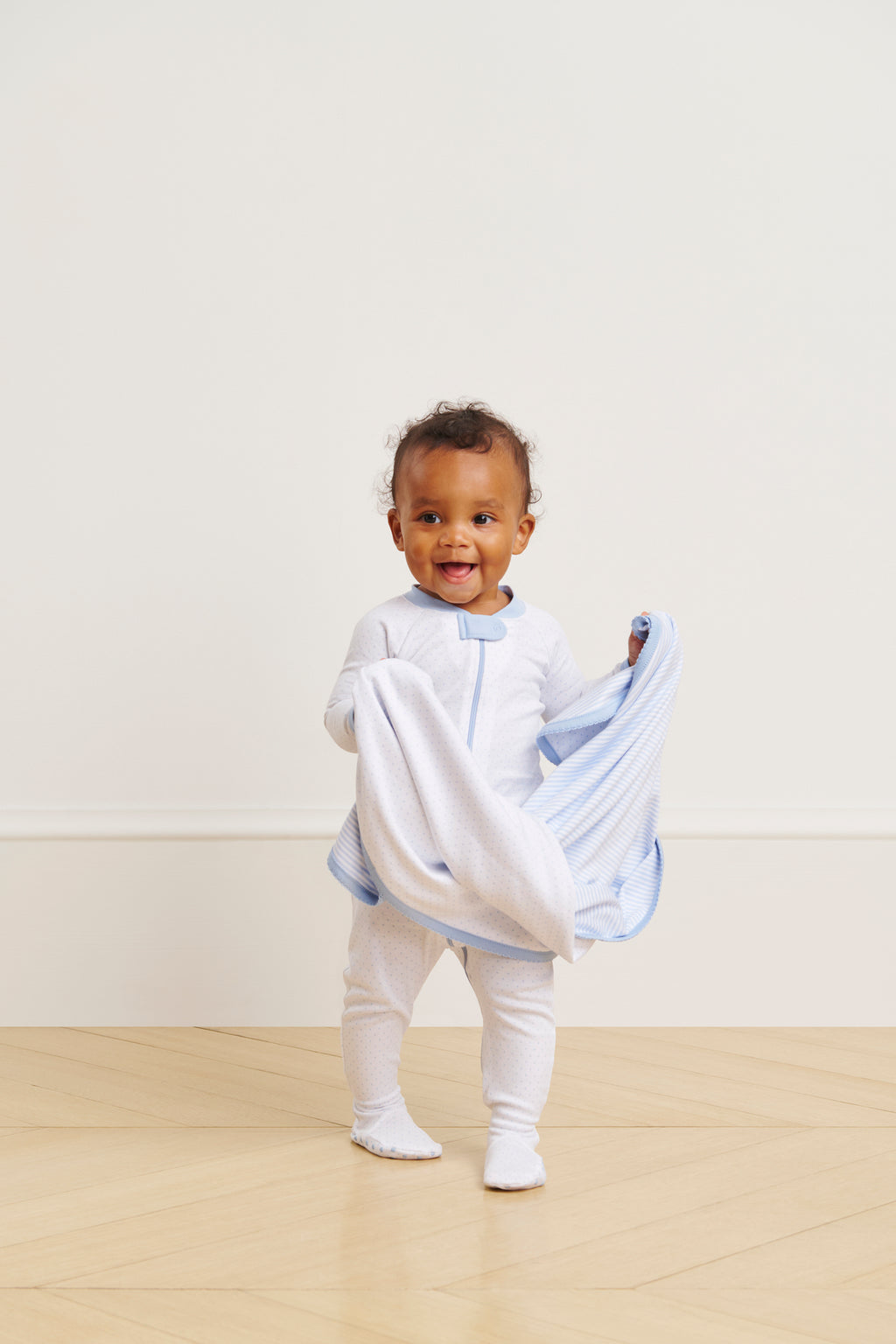 A smiling baby stands on a wooden floor, holding a soft light blue blanket, wearing the Baby Sleeper in French Blue Pindot. The background is a plain light-colored wall.