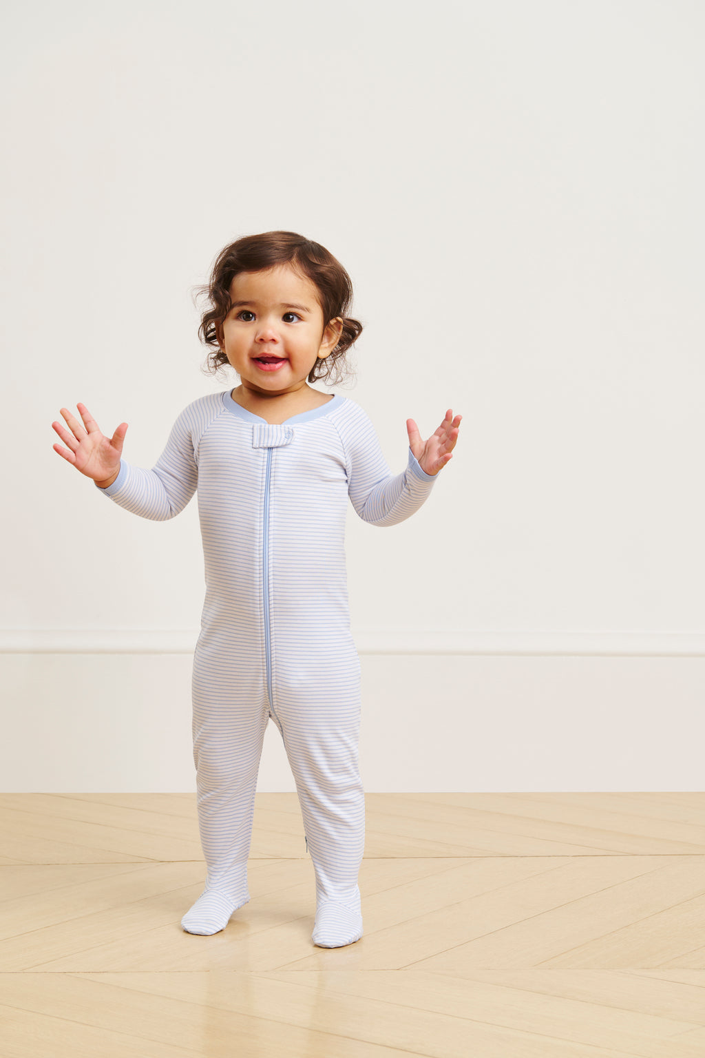 A toddler with a cheerful expression stands on a wooden floor wearing the Baby Sleeper in French Blue; the background is plain and light-colored.