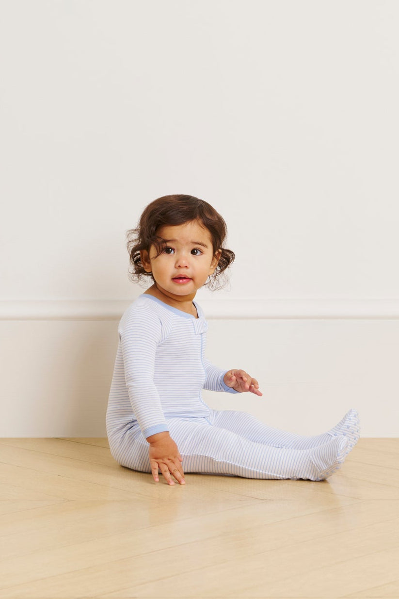 A toddler sits on a light wooden floor against a pale wall, wearing the Baby Sleeper in French Blue. The child looks slightly to the side with one hand resting on the floor.