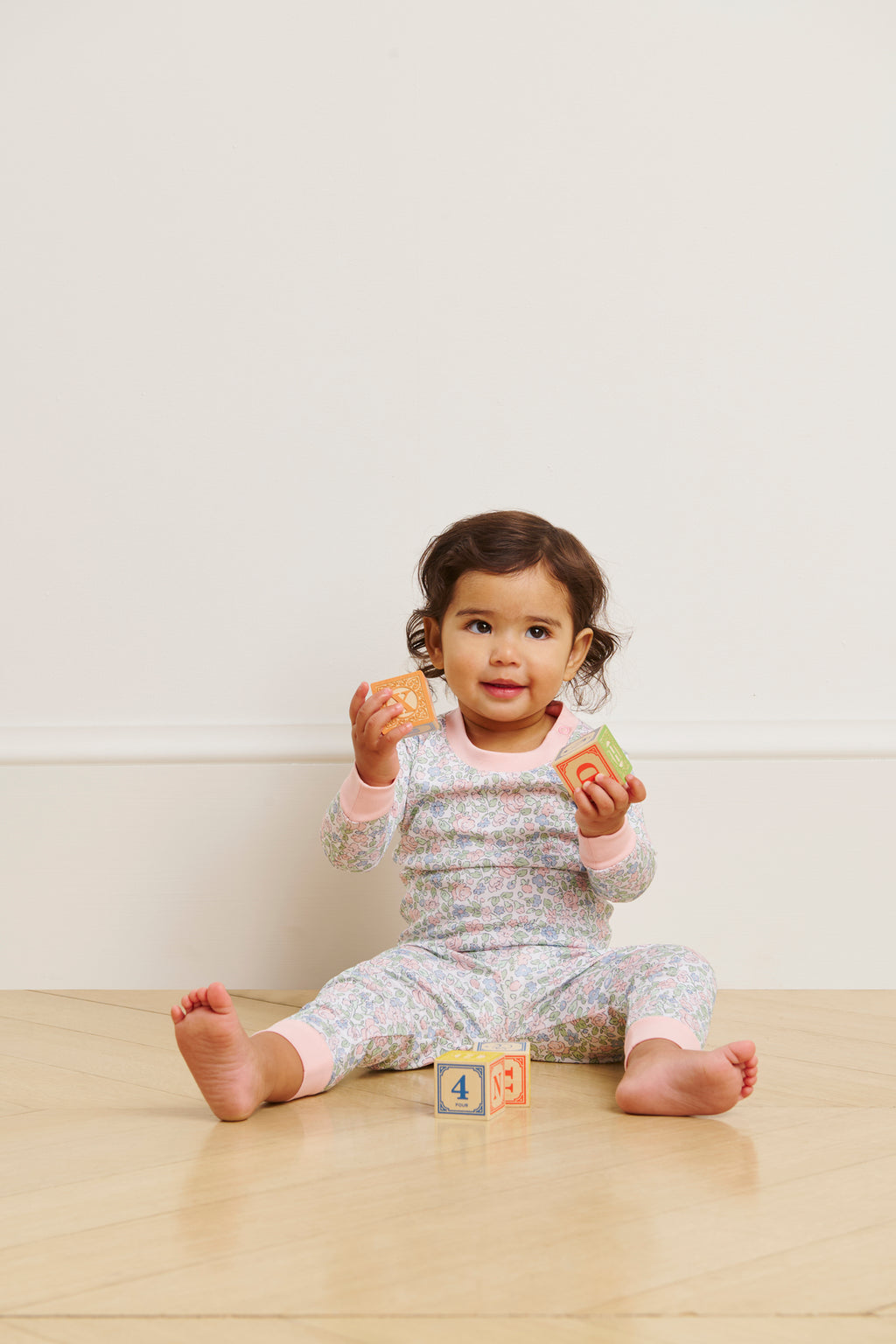 A young child in a Baby Long-Long Set in English Rose Elizabeth Floral sits on a wooden floor, holding colorful alphabet blocks, with more blocks scattered nearby against a plain white wall.