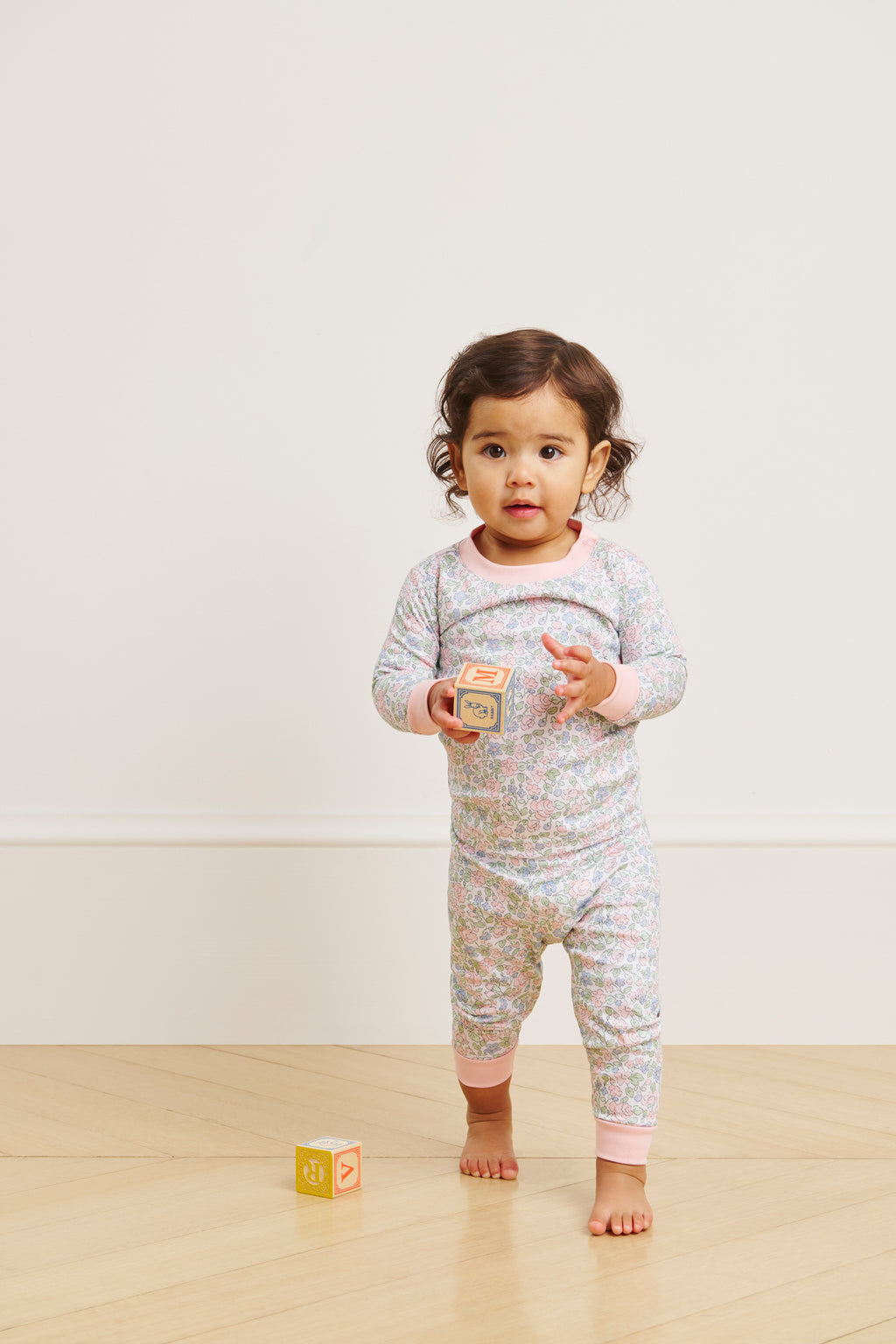 A toddler in a Baby Long-Long Set in English Rose Elizabeth Floral stands on a wooden floor, holding a wooden block with another nearby. The background features a plain, light-colored wall.