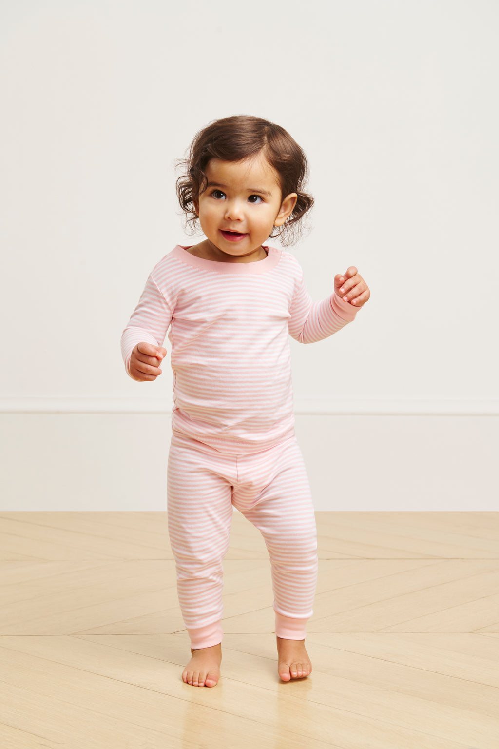 A cheerful toddler stands on a wooden floor, wearing the Baby Long-Long Set in English Rose Stripe with light pink and white stripes.