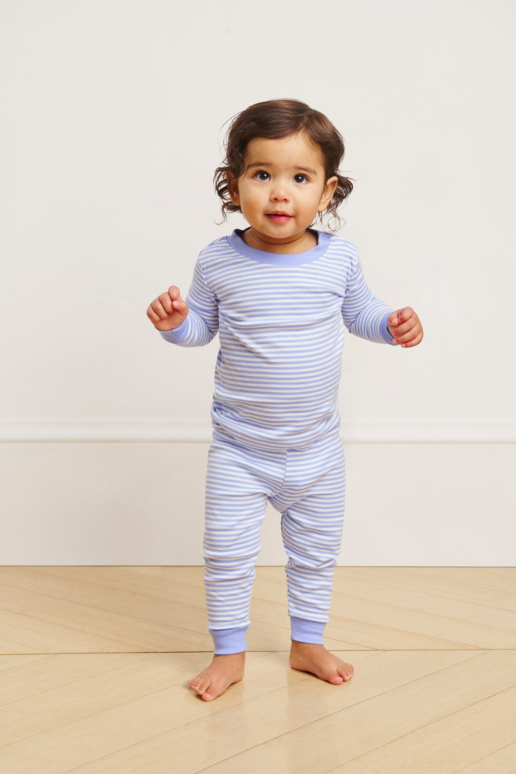 A young child stands on a wooden floor, smiling and looking up while wearing the Baby Long-Long Set in Hydrangea.