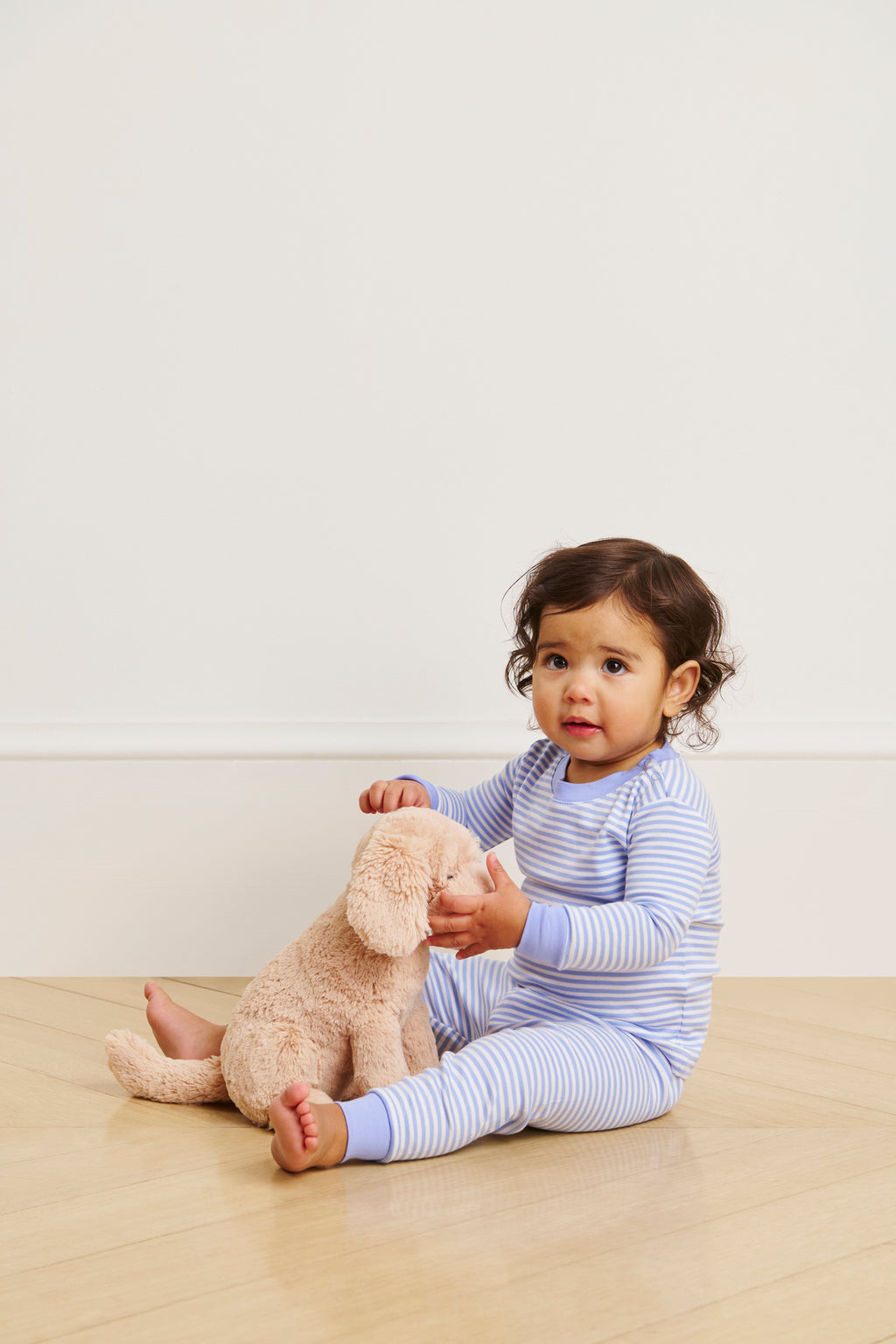 A young child in a Baby Long-Long Set in Hydrangea sits on a wooden floor, holding a plush beige stuffed animal. Made from soft Pima Cotton, the outfit stands out against the plain, light-colored wall.