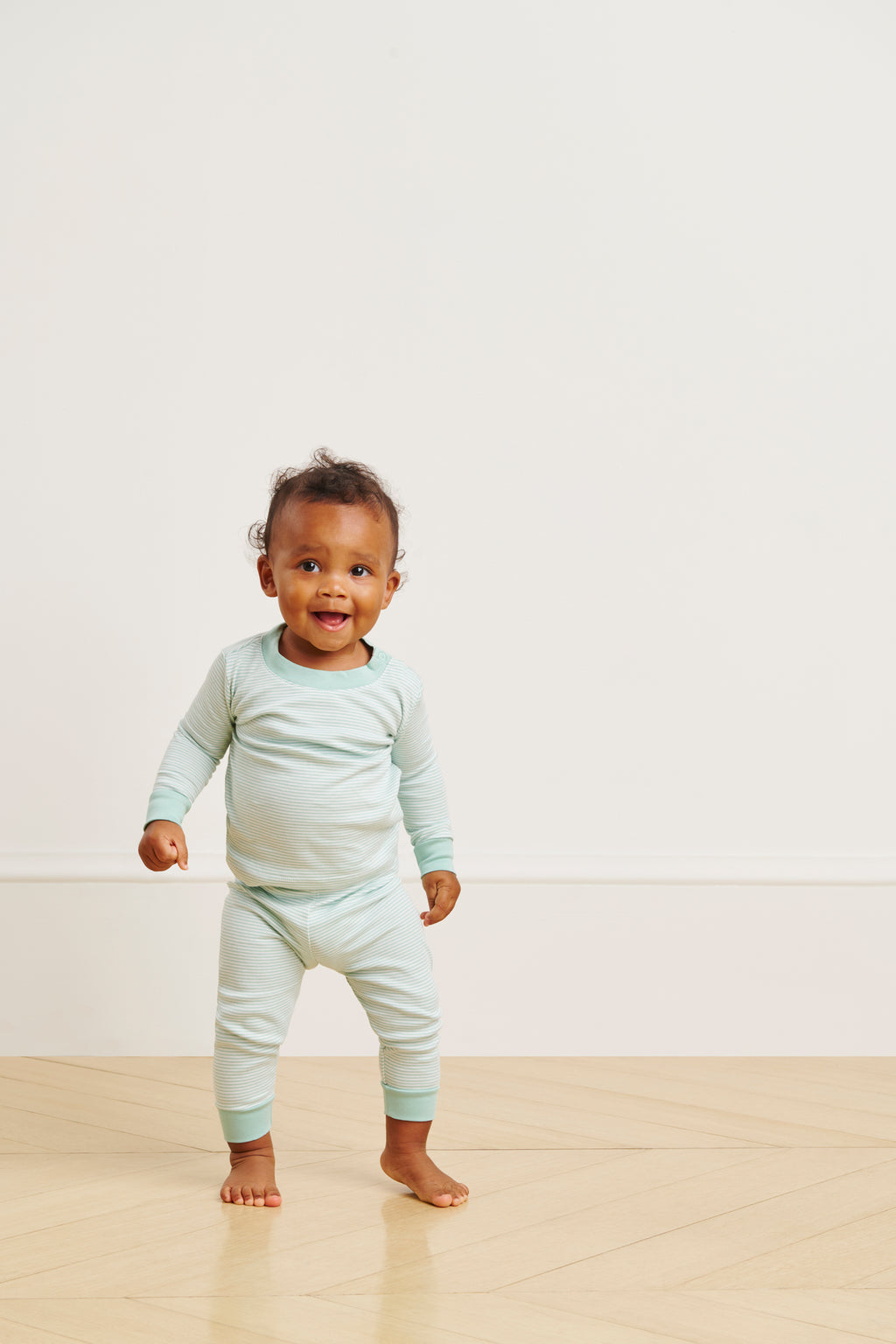 A smiling toddler wearing a Parisian Green Baby Long-Long Set stands on a wooden floor against a plain, light-colored wall.