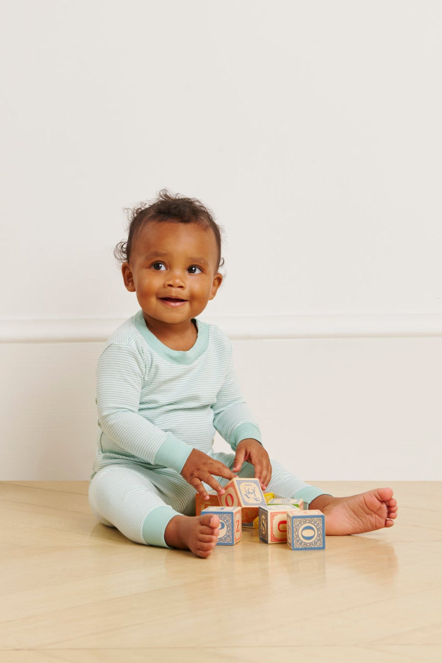 A baby in a Baby Long-Long Set in Parisian Green smiles while playing with colorful alphabet blocks on a wooden floor, against a simple background with a light wall and white trim.