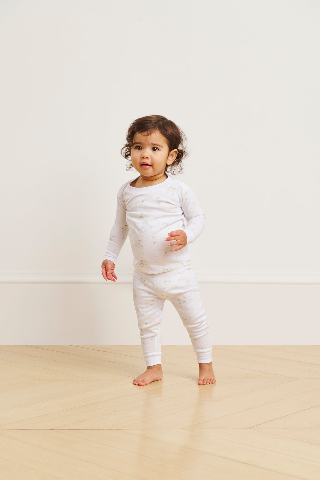 A toddler wearing the Baby Long-Long Set in Sleepy Sheep, featuring a white Pima Cotton long-sleeve top and matching pants, stands on a light wooden floor with a plain, light background.
