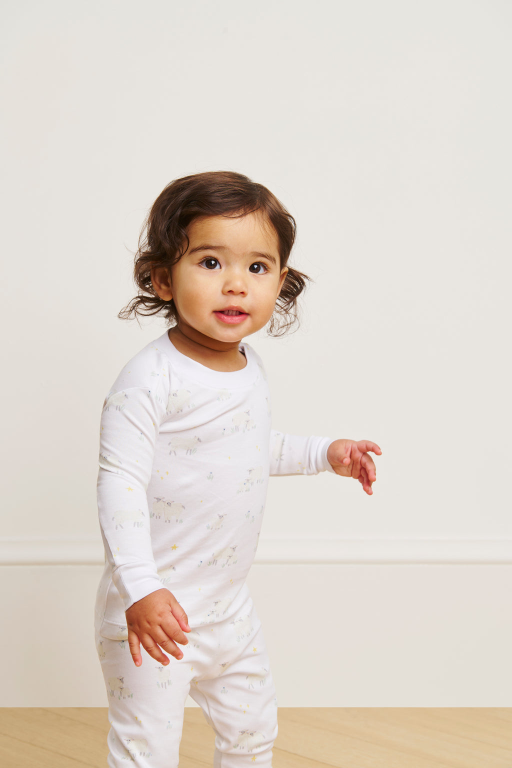 A toddler wearing the Baby Long-Long Set in Sleepy Sheep, made from soft Pima cotton with small patterns, stands indoors on a light wood floor against a plain, light wall, looking slightly to the side.