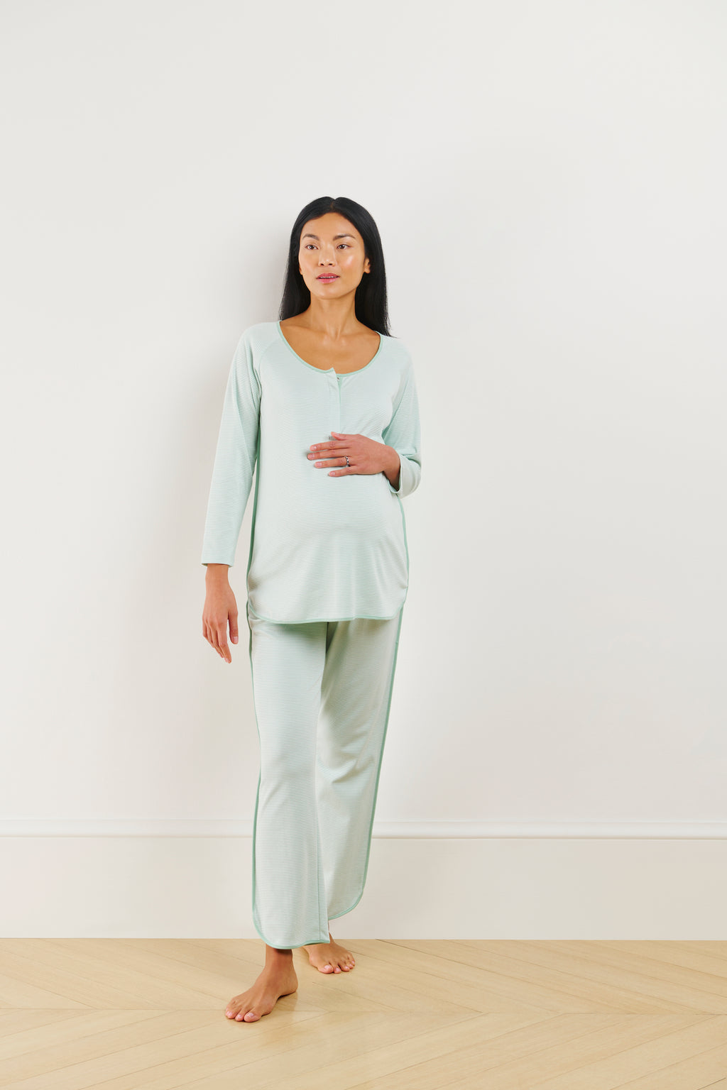A woman stands indoors against a plain white wall, wearing the Pima Maternity Long-Long Set in Parisian Green, made from 100% Pima cotton. She rests one hand on her stomach and gazes ahead with a neutral expression.