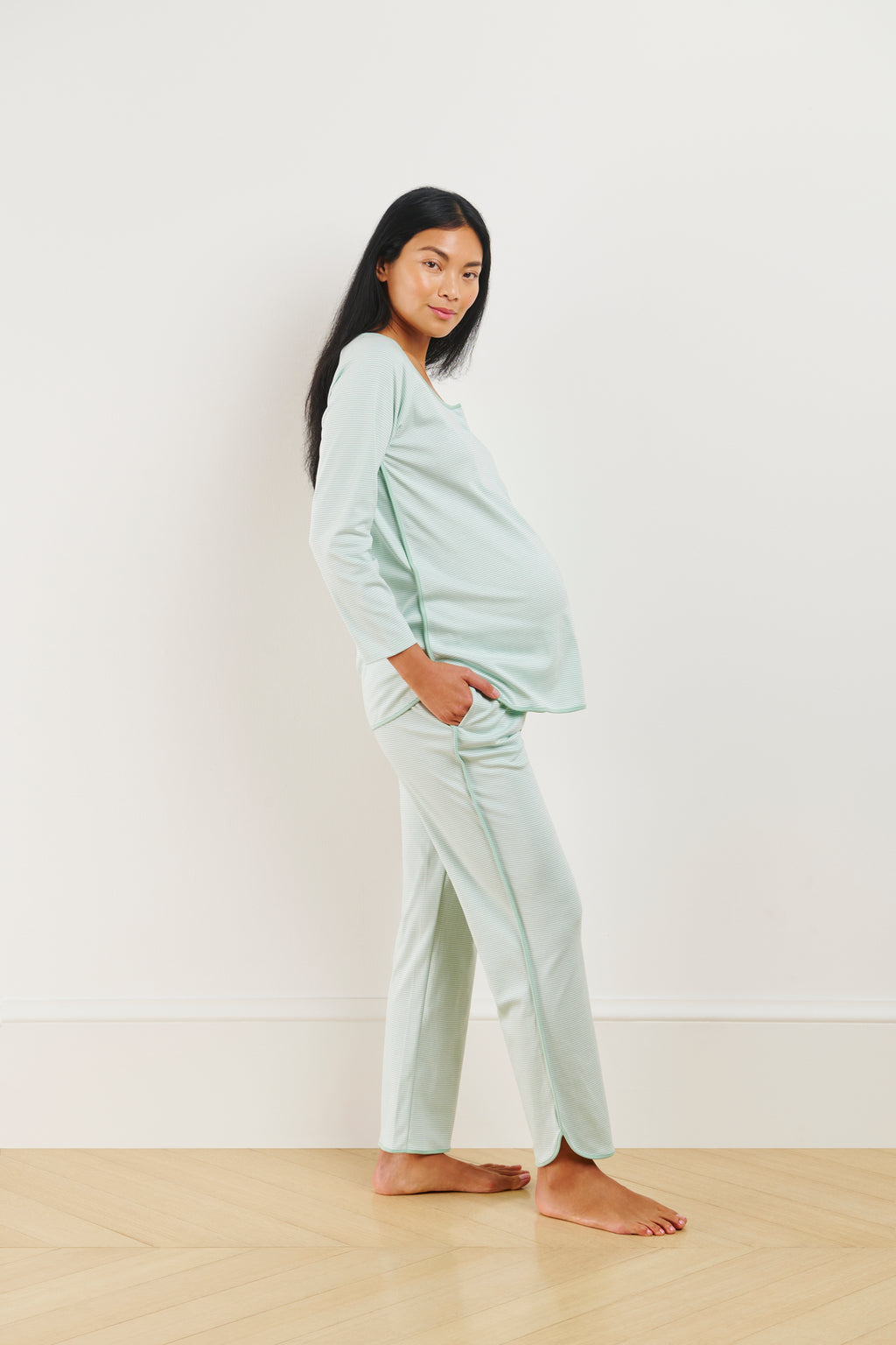 A pregnant woman stands on a wooden floor, wearing the Pima Maternity Long-Long Set in Parisian Green, made of 100% Pima cotton, with one hand in her pocket and looking calmly at the camera against a plain white wall.