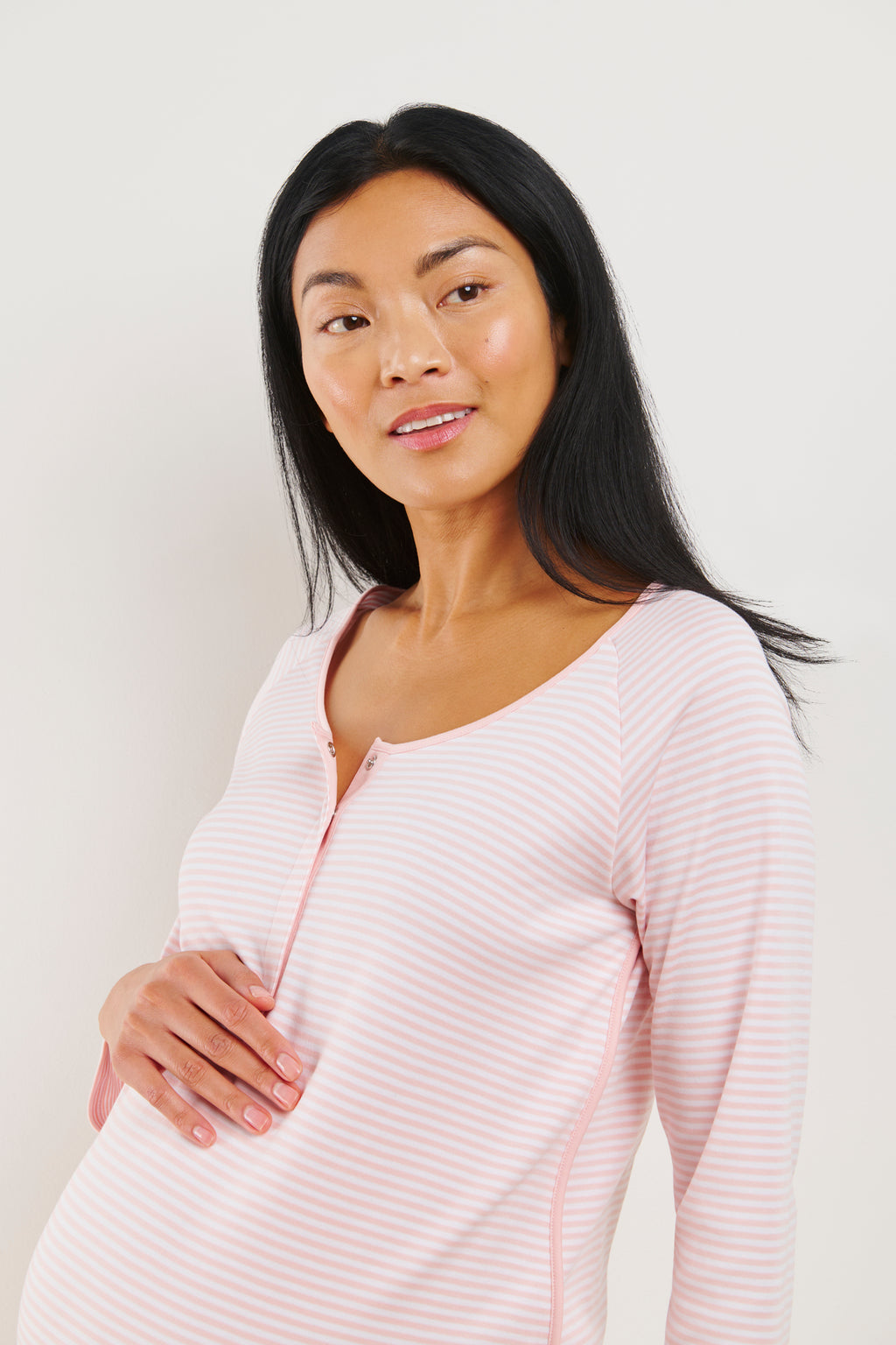A woman smiles softly and gently touches her belly while wearing the Pima Maternity Long Sleeve Nightgown in English Rose Stripe, standing against a plain light background.