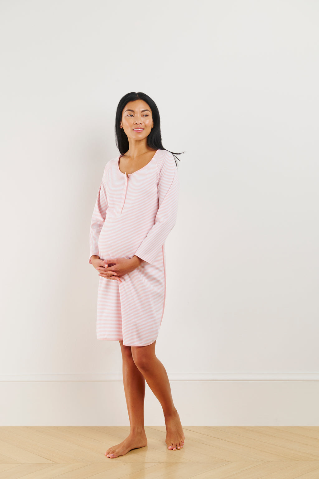 A pregnant woman stands on a wooden floor, wearing the Pima Maternity Long Sleeve Nightgown in English Rose Stripe. She gently holds her belly and gazes to the side against a plain white wall.