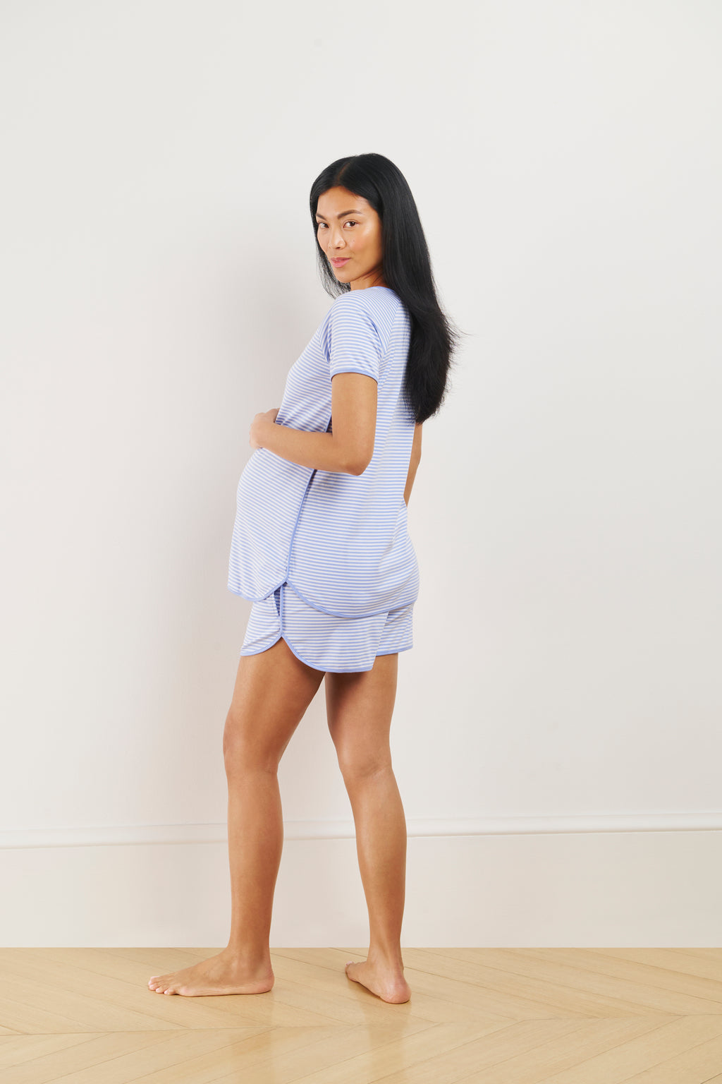 A woman stands barefoot on a wooden floor, wearing the Pima Maternity Shorts Set in Hydrangea, facing slightly to the side and looking at the camera against a plain light background.