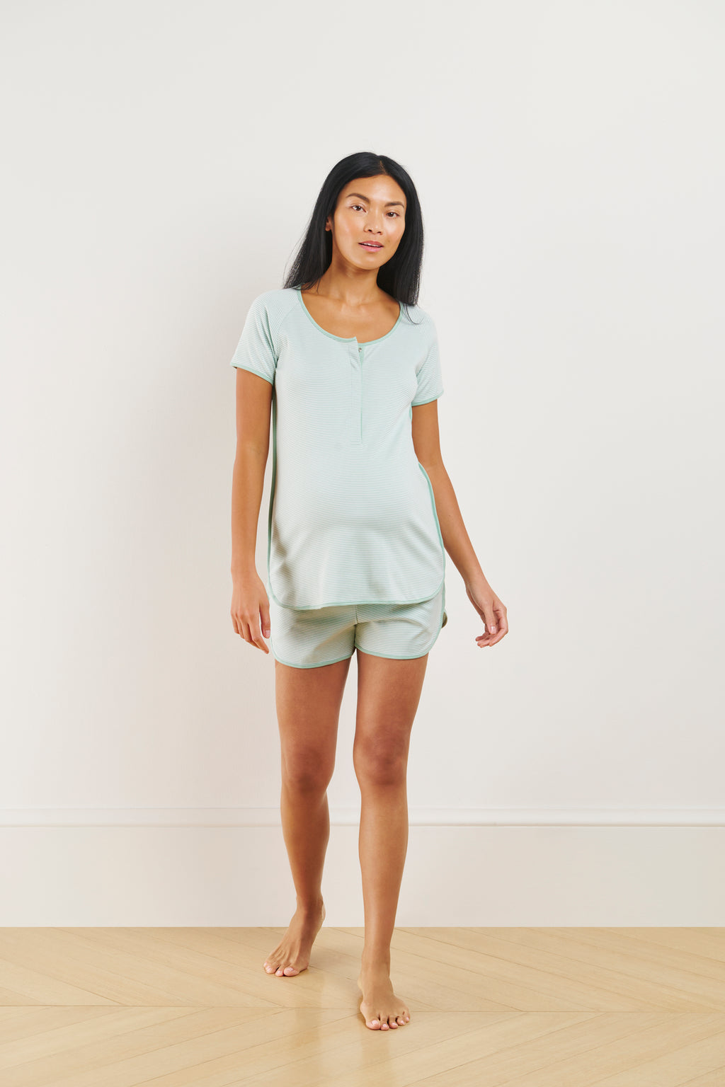 A woman stands barefoot on a wooden floor against a white wall, wearing the Pima Maternity Shorts Set in Parisian Green.